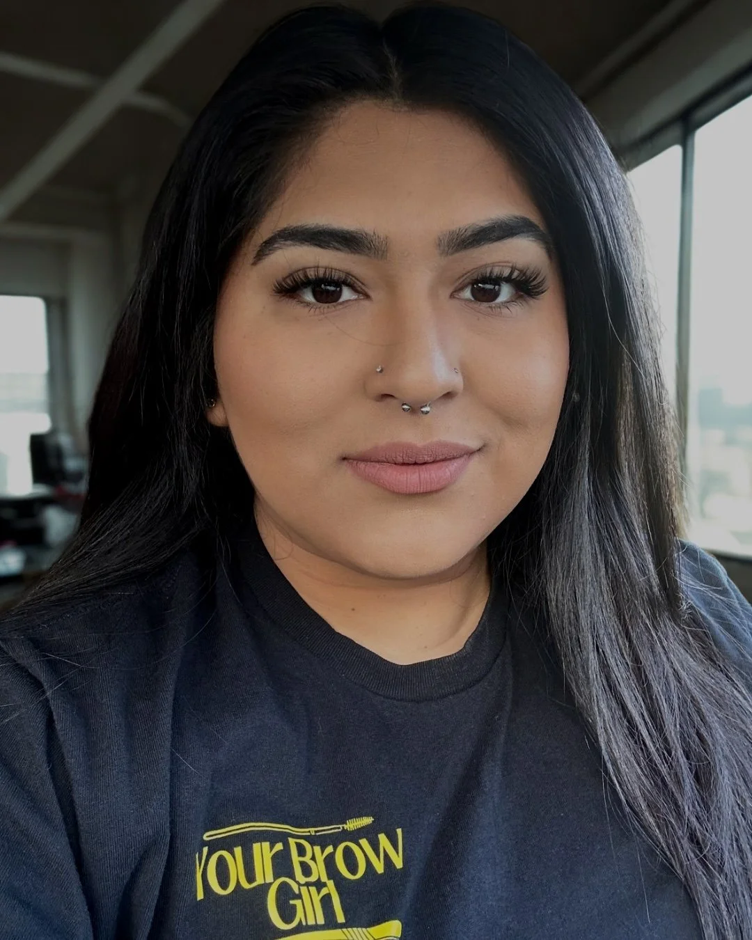 A woman with long dark hair wearing makeup and a septum piercing, smiling at the camera, in an indoor setting with windows in the background.