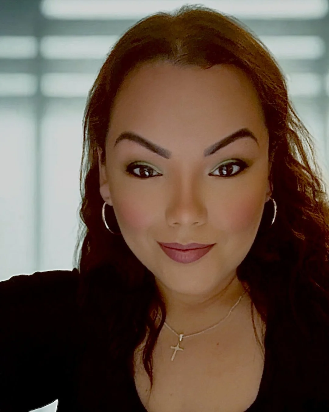 Close-up of a woman with long, curly brown hair, wearing hoop earrings and a cross necklace, smiling at the camera.