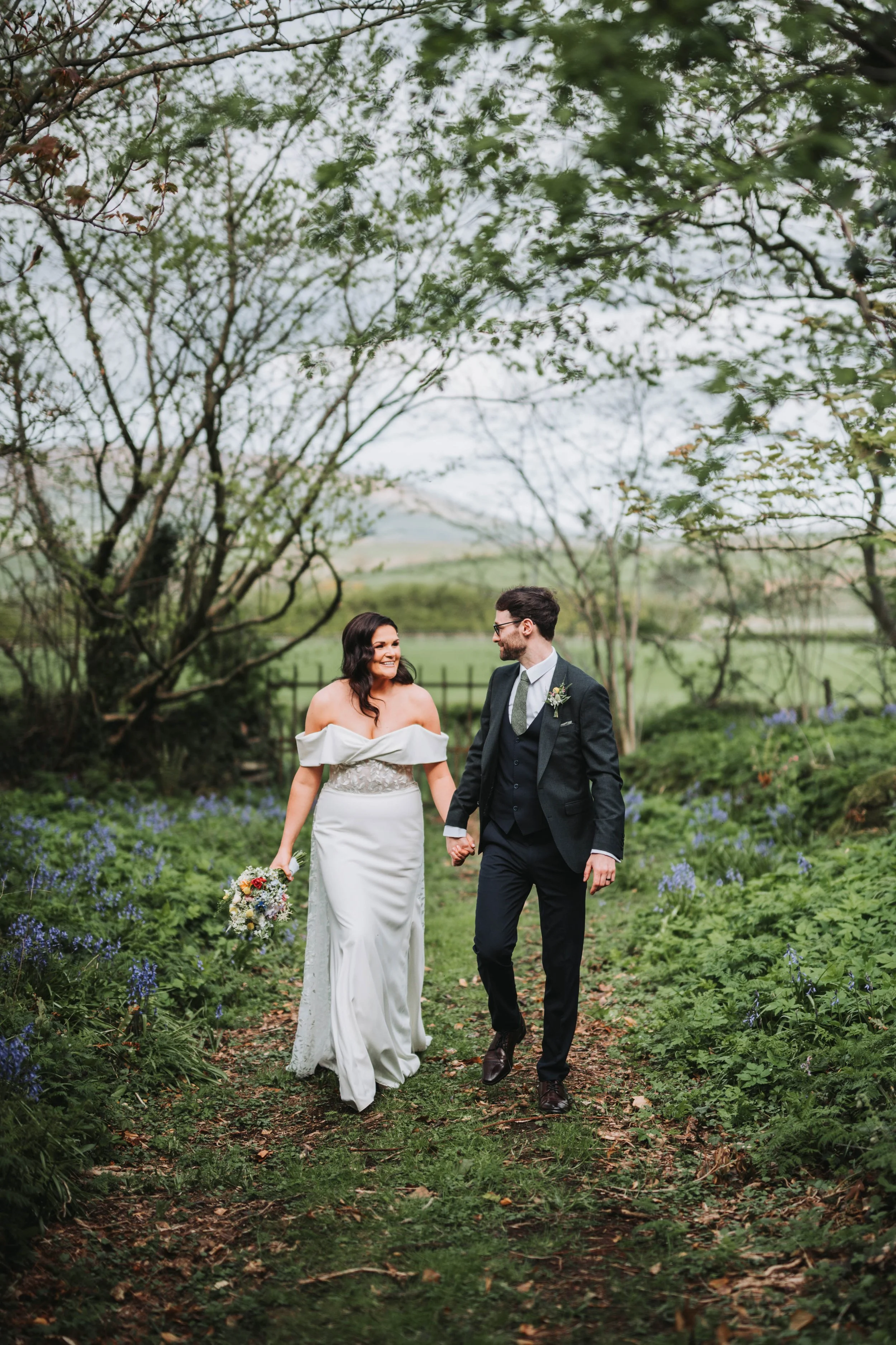 A newlywed couple walking hand in hand through a lush garden or park, surrounded by green foliage and purple flowers, with trees in the background.