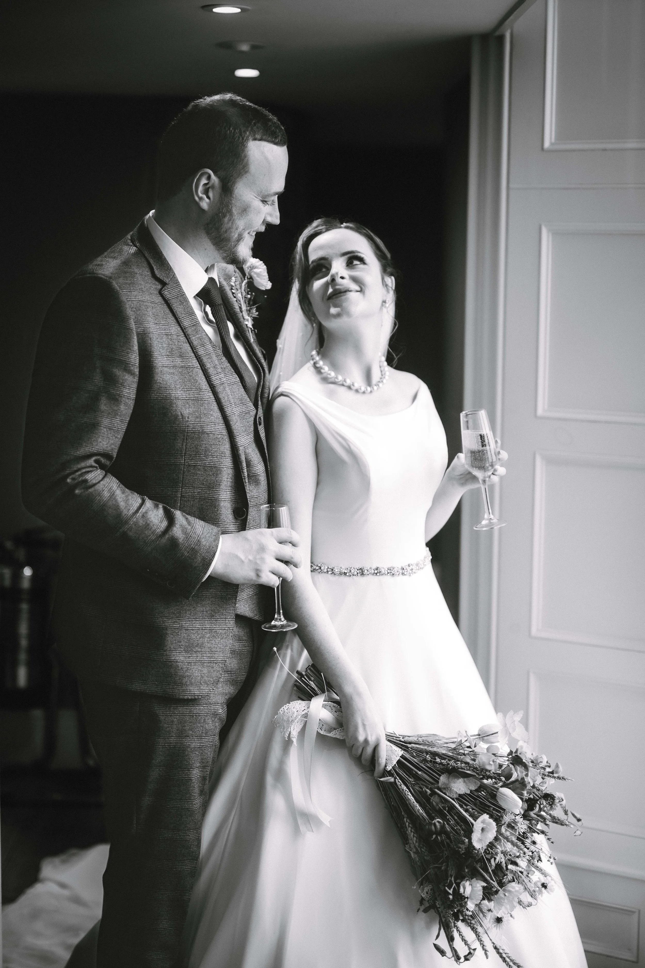 Black and white photo of a bride and groom at their wedding. The groom is wearing a checked suit and holding a champagne glass. The bride, in a satin wedding dress with pearl necklace and belt, holds a bouquet and a champagne glass. They are smiling 