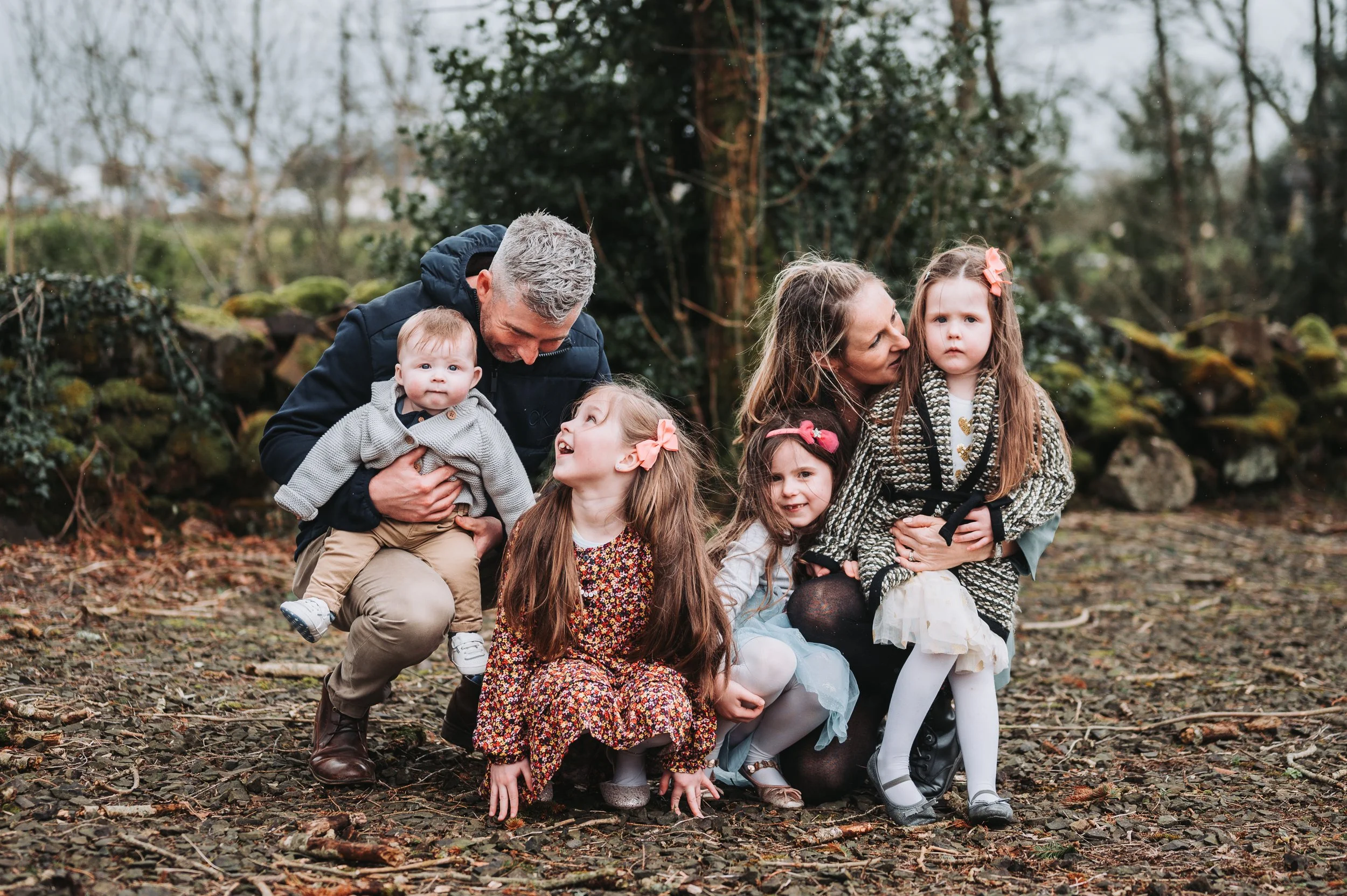 Family of six outdoors in a natural setting with trees and rocks, including two adults and four children, some smiling and some with neutral expressions.