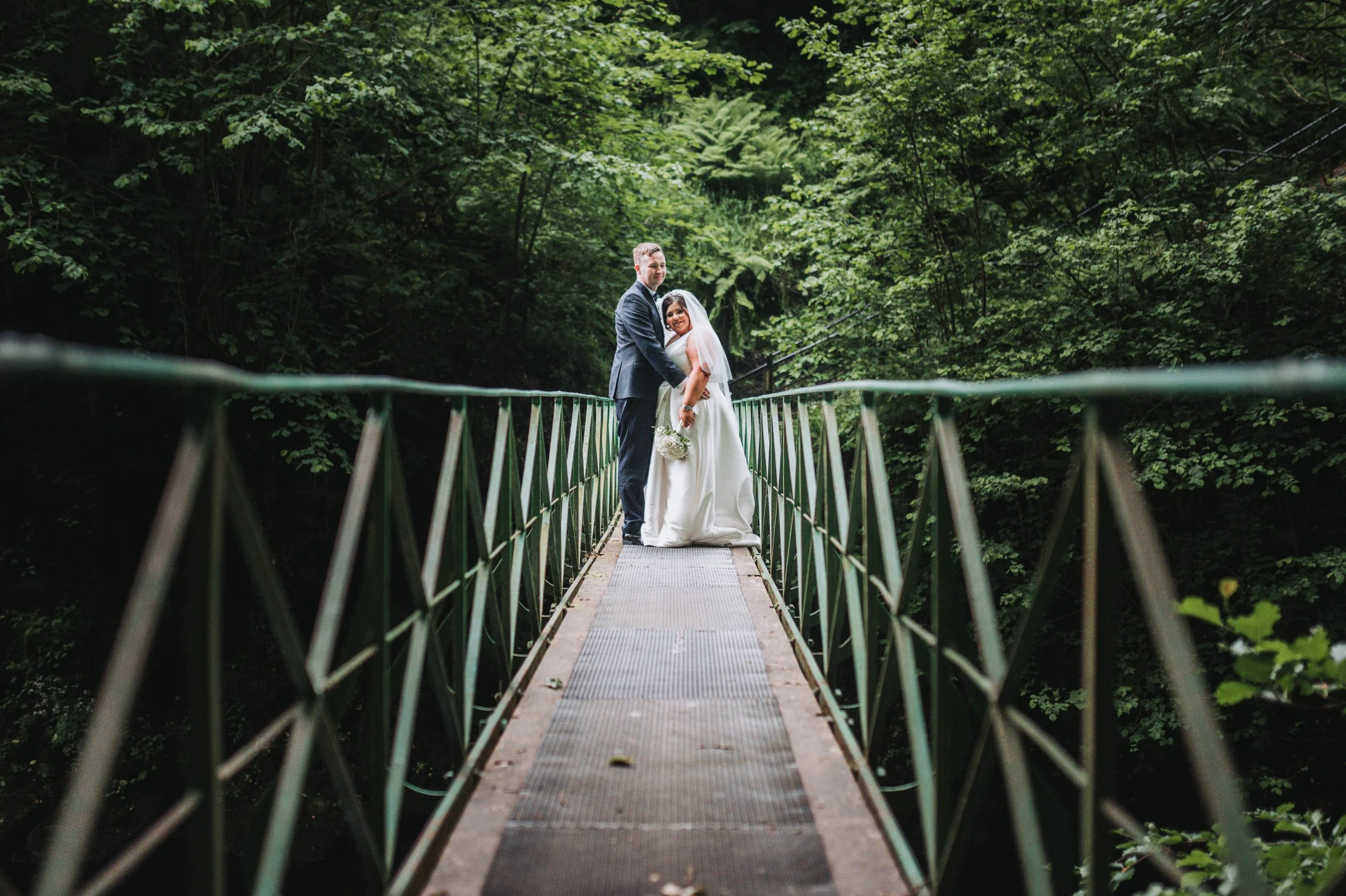 Bride and groom standing on a narrow metal bridge surrounded by lush green trees.