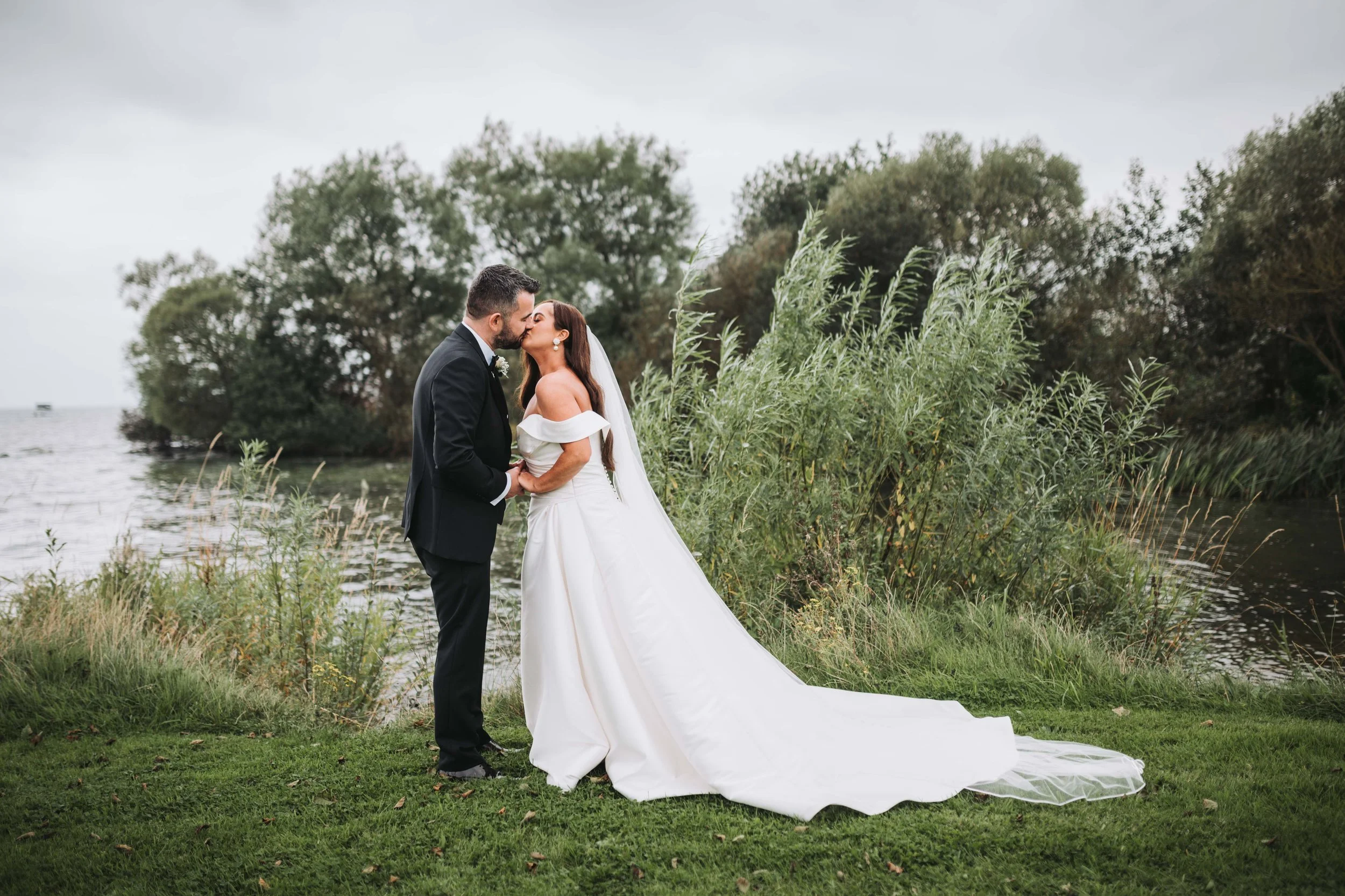 A bride and groom sharing a kiss outdoors near a body of water with lush green trees and grass in the background.