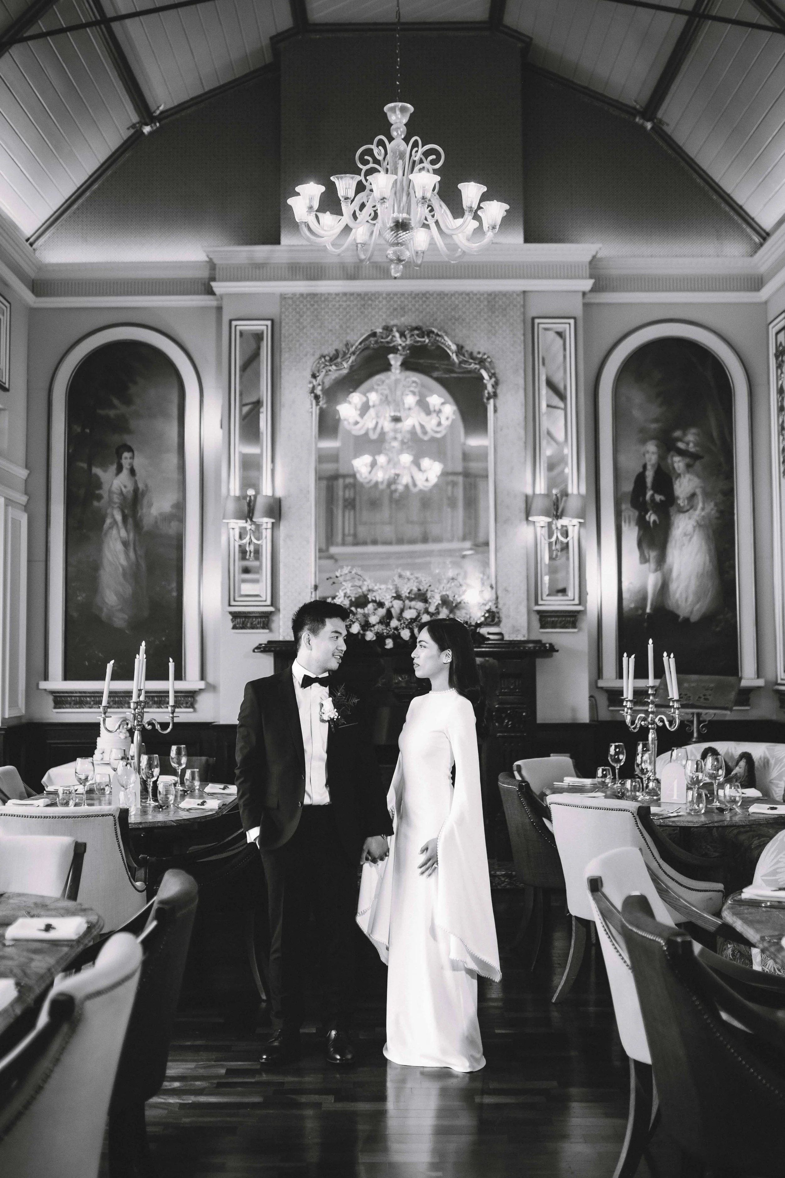 A black and white photo of a bride and groom standing close together in an elegant, ornately decorated dining room with chandeliers, portraits, and a flower bouquet in the background.