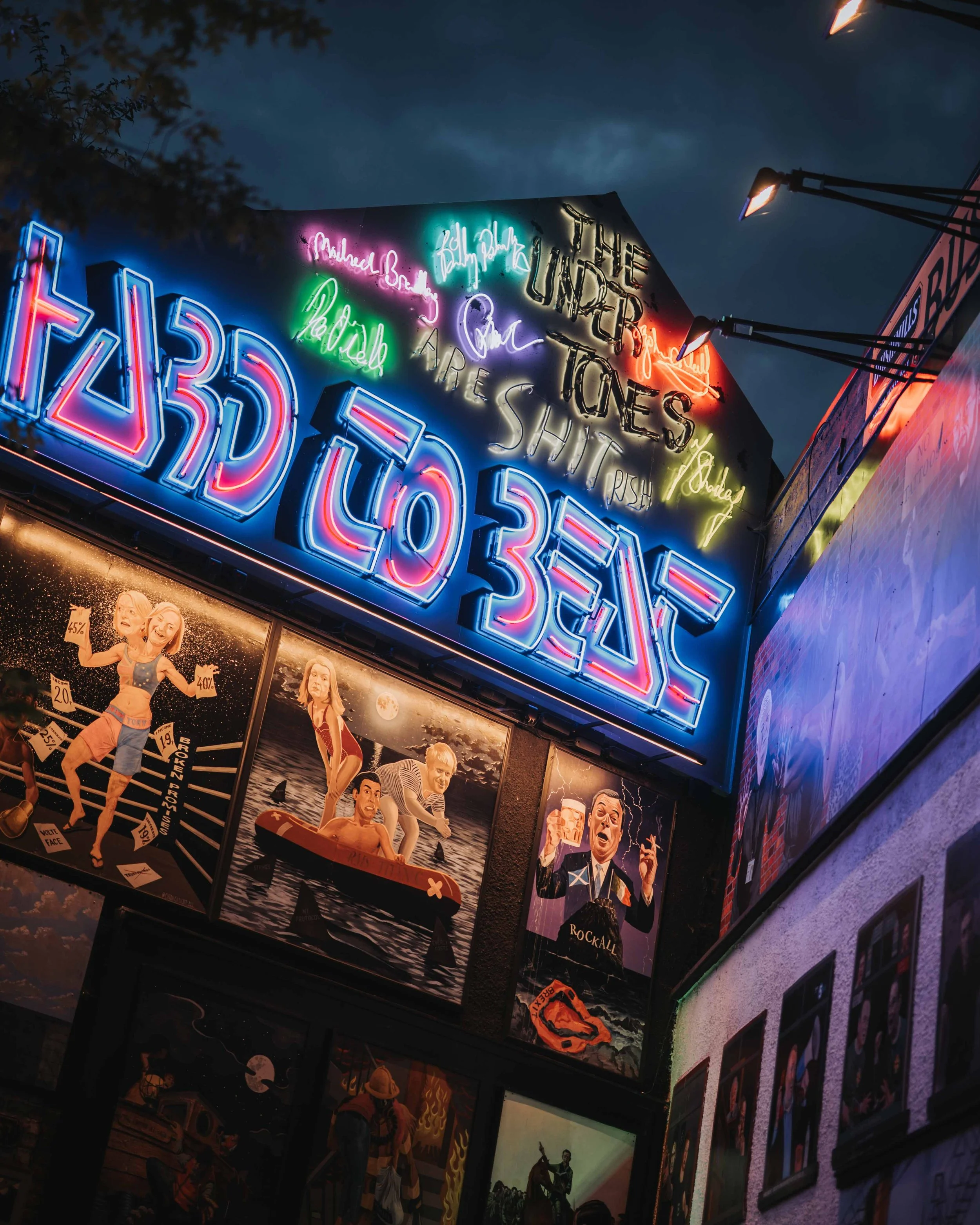 Neon sign reading 'Prague' with various colorful lights, surrounding art posters including surfing and newsvendor illustrations, and a cloudy sky.