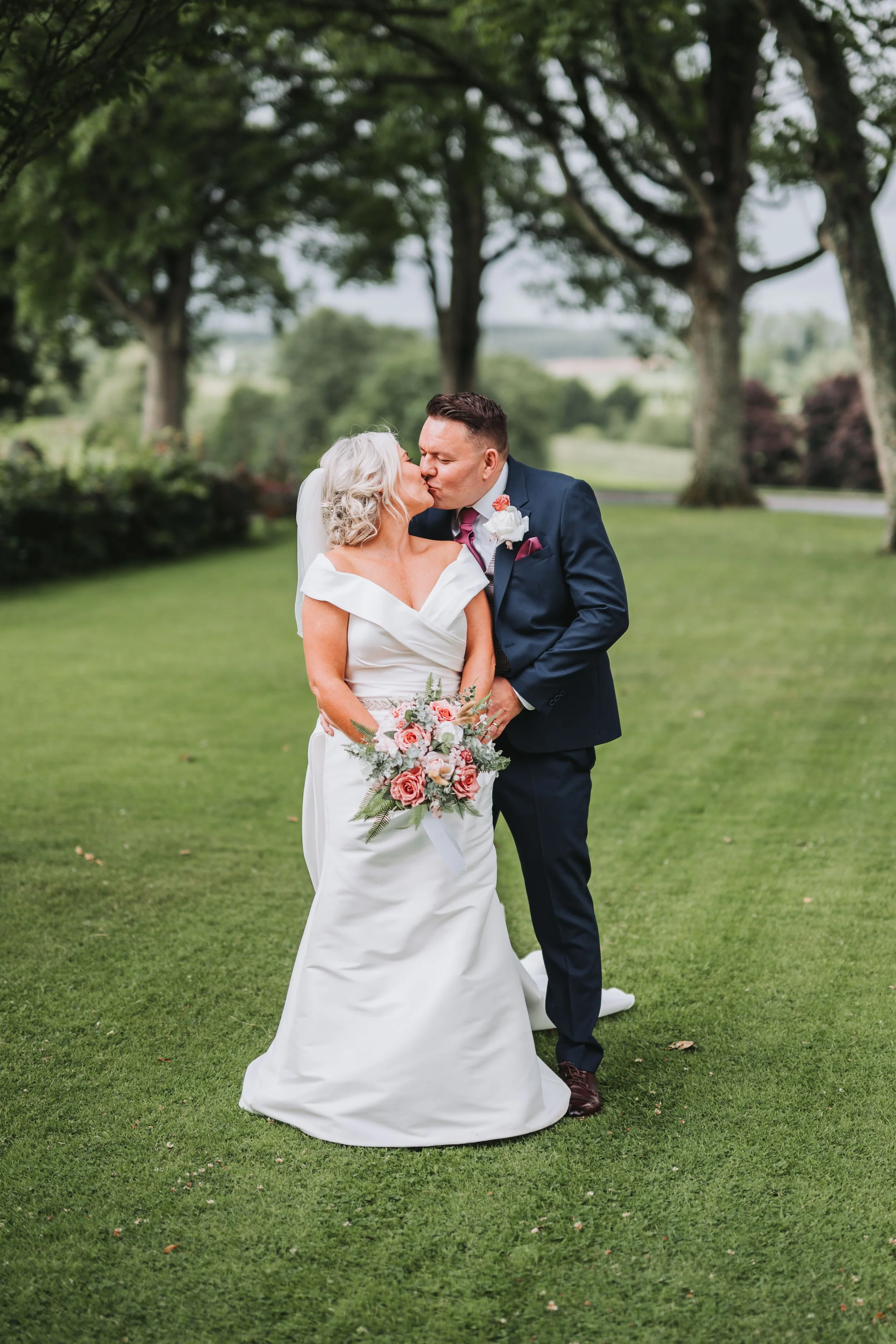 A bride and groom sharing a kiss outdoors on a green lawn with trees in the background, the bride holding a bouquet of pink and white flowers