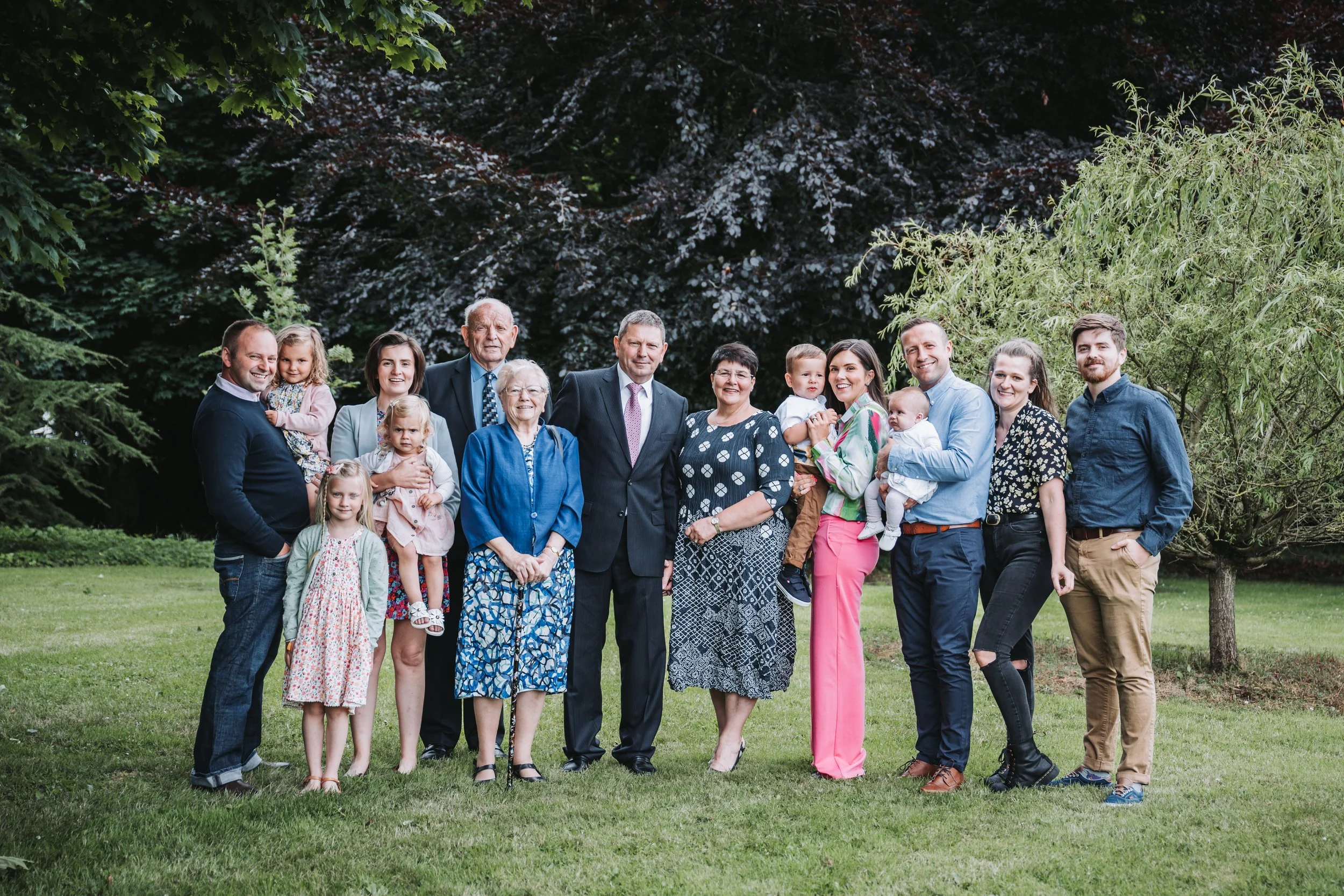 A large multigenerational family photo taken outdoors in a grassy area with trees in the background. The group includes children, adults, and elderly, all smiling and posing together.