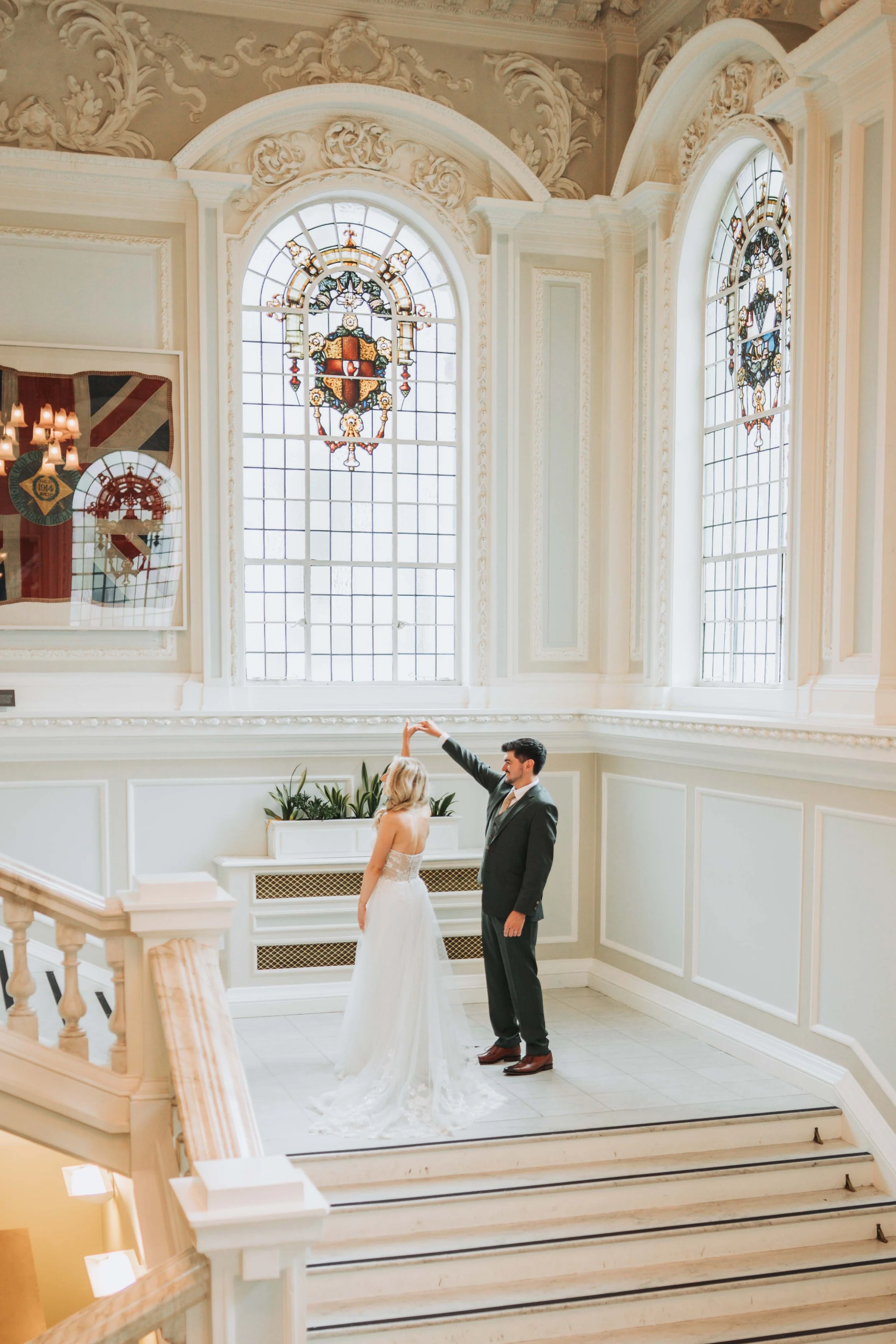 A bride and groom dancing in a grand, ornate room with large stained glass windows, decorative molding, and a staircase.