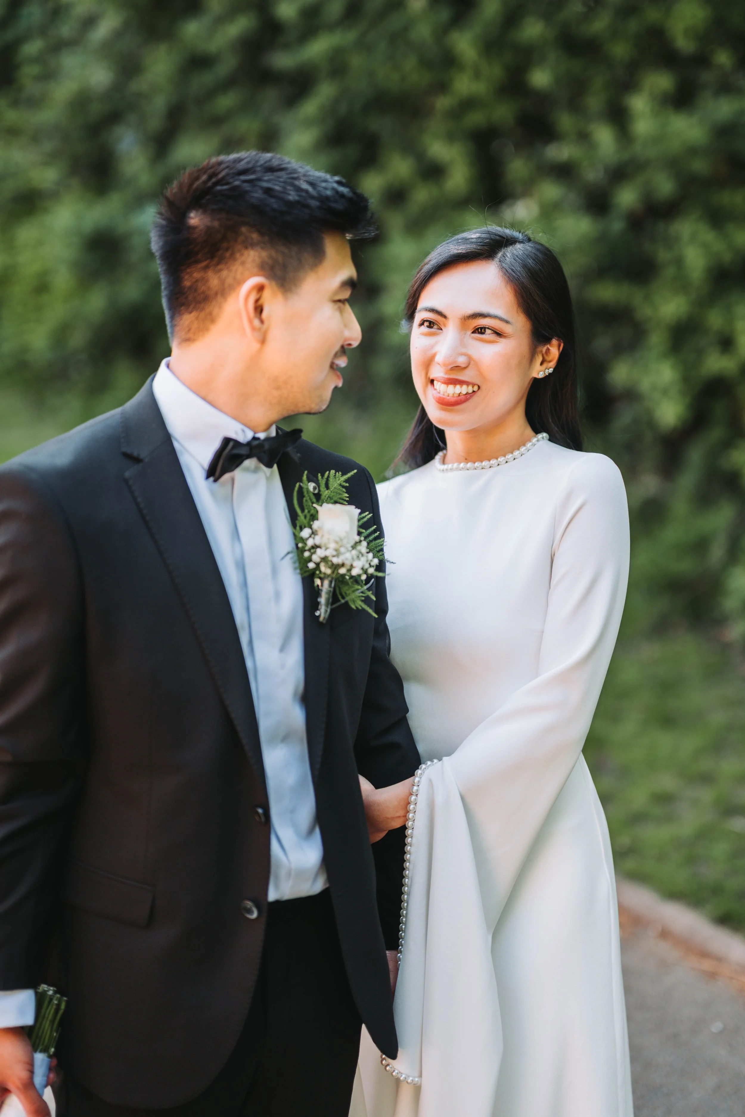 A bride and groom standing outdoors on their wedding day, gazing at each other with a background of green trees.