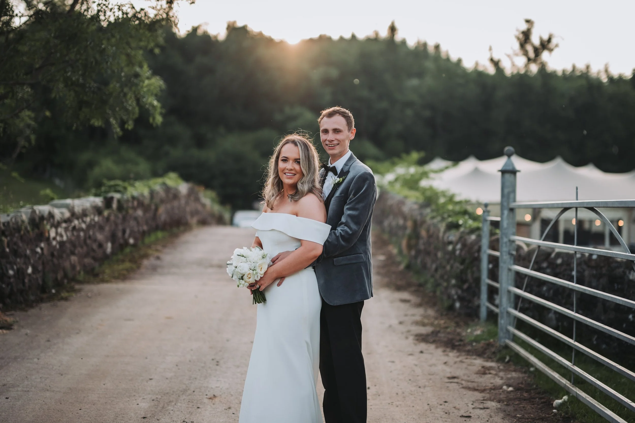 A bride and groom standing together on a dirt path outdoors during sunset, with trees in the background and a metal gate on the right, both smiling at the camera. The bride holds a bouquet of white roses, and the groom wears a gray tuxedo with a blac