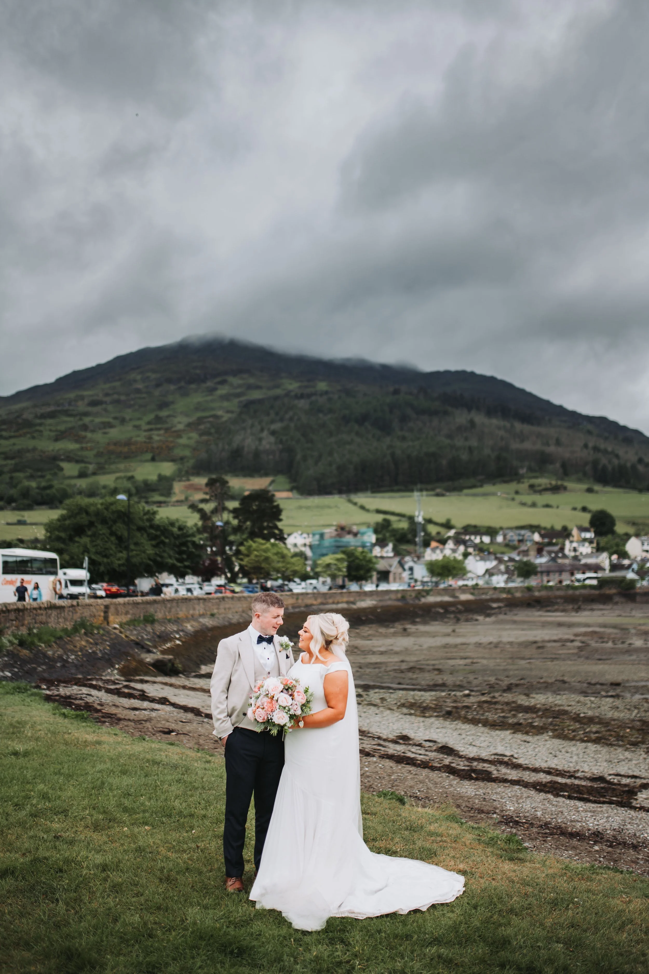 Bride and groom standing on grass with a mountain and cloudy sky in the background. The bride is holding a bouquet, and they are gazing at each other.