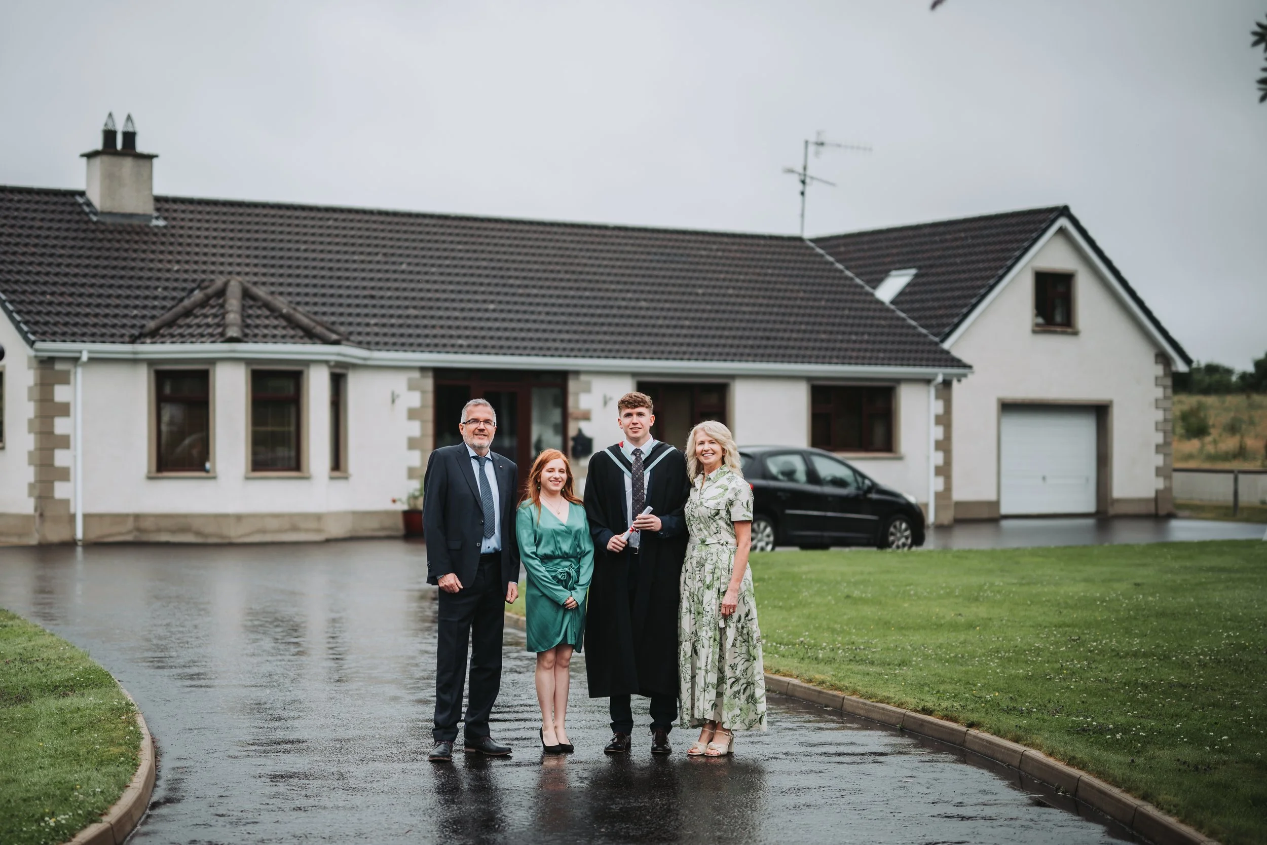 Family celebrating graduation outdoors on a rainy day, standing on wet driveway in front of a house.