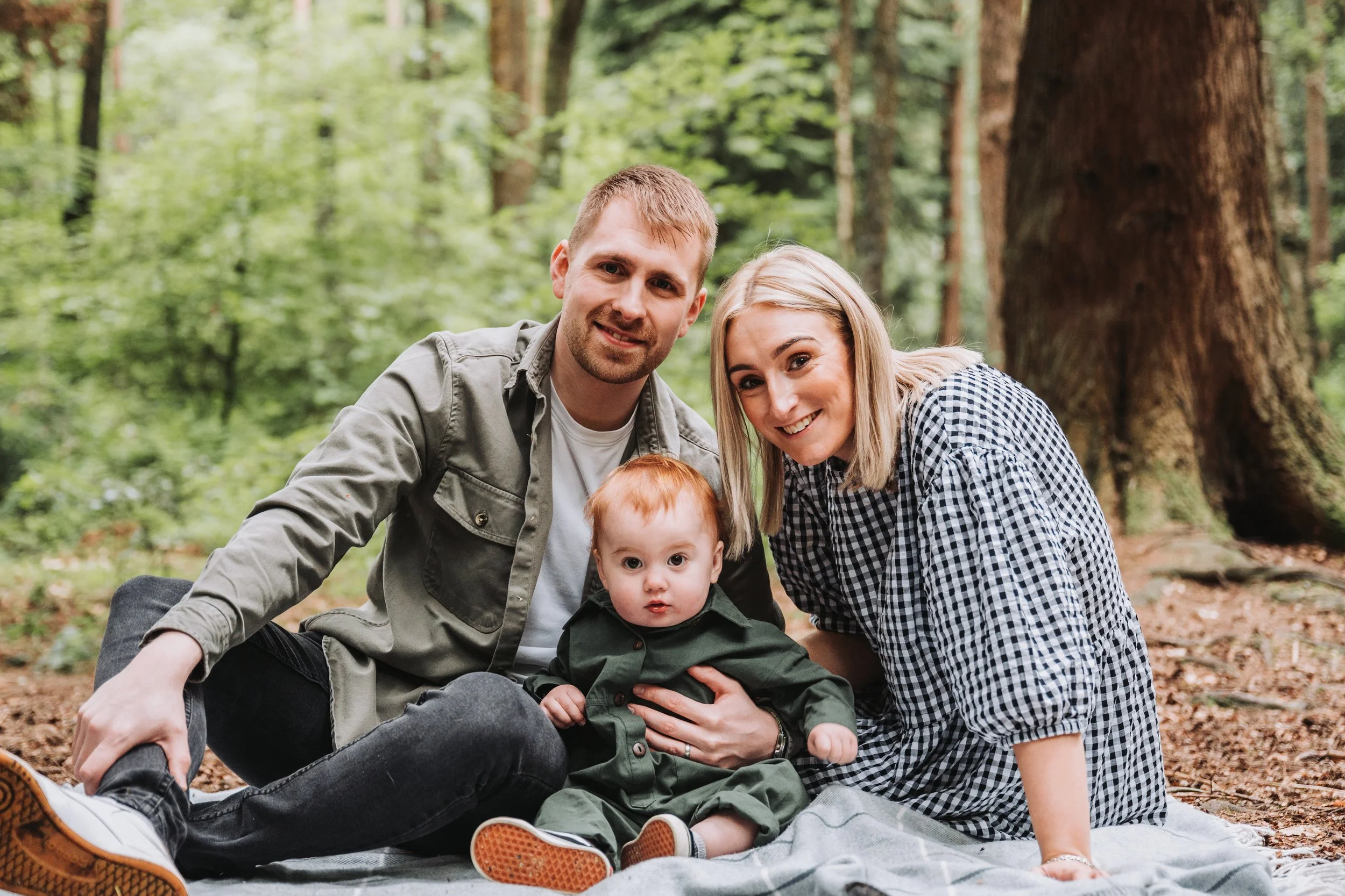 A family of three sitting on a blanket in a forest, smiling at the camera. The father, mother, and young child are surrounded by tall trees and green foliage.