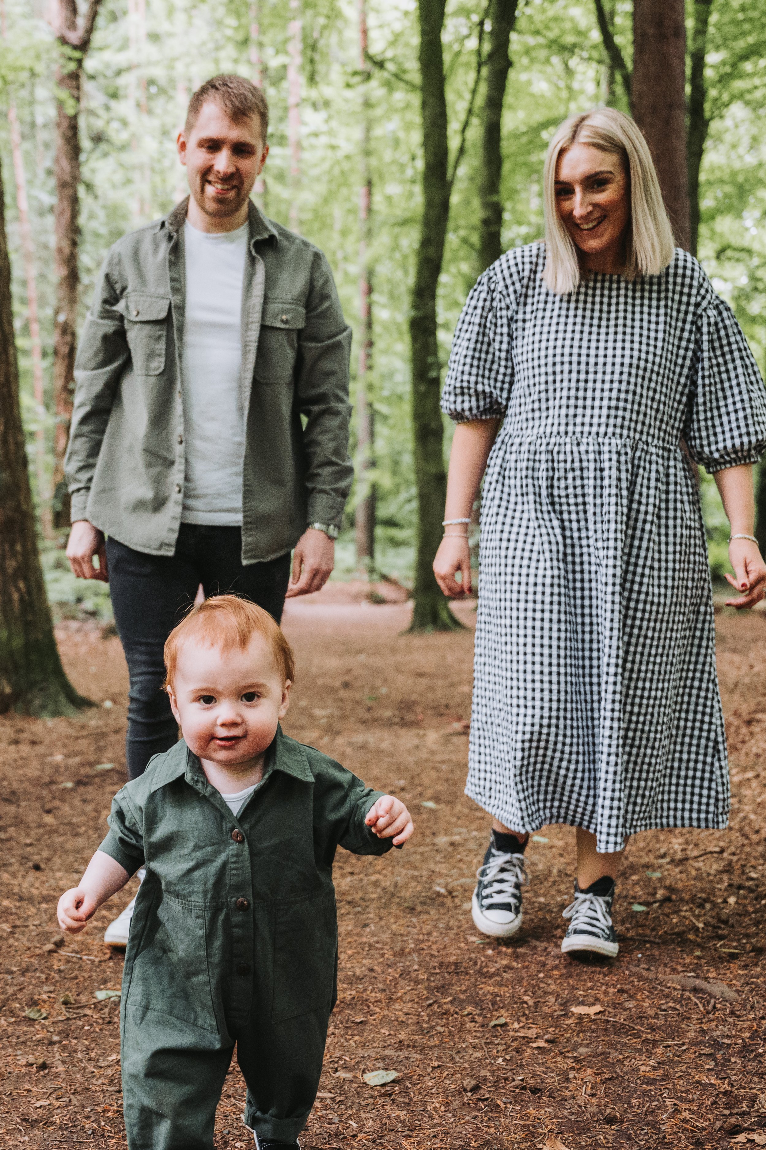 A family of three walking on a wooded trail, with a young toddler in the foreground and two adults walking behind him, surrounded by tall green trees.
