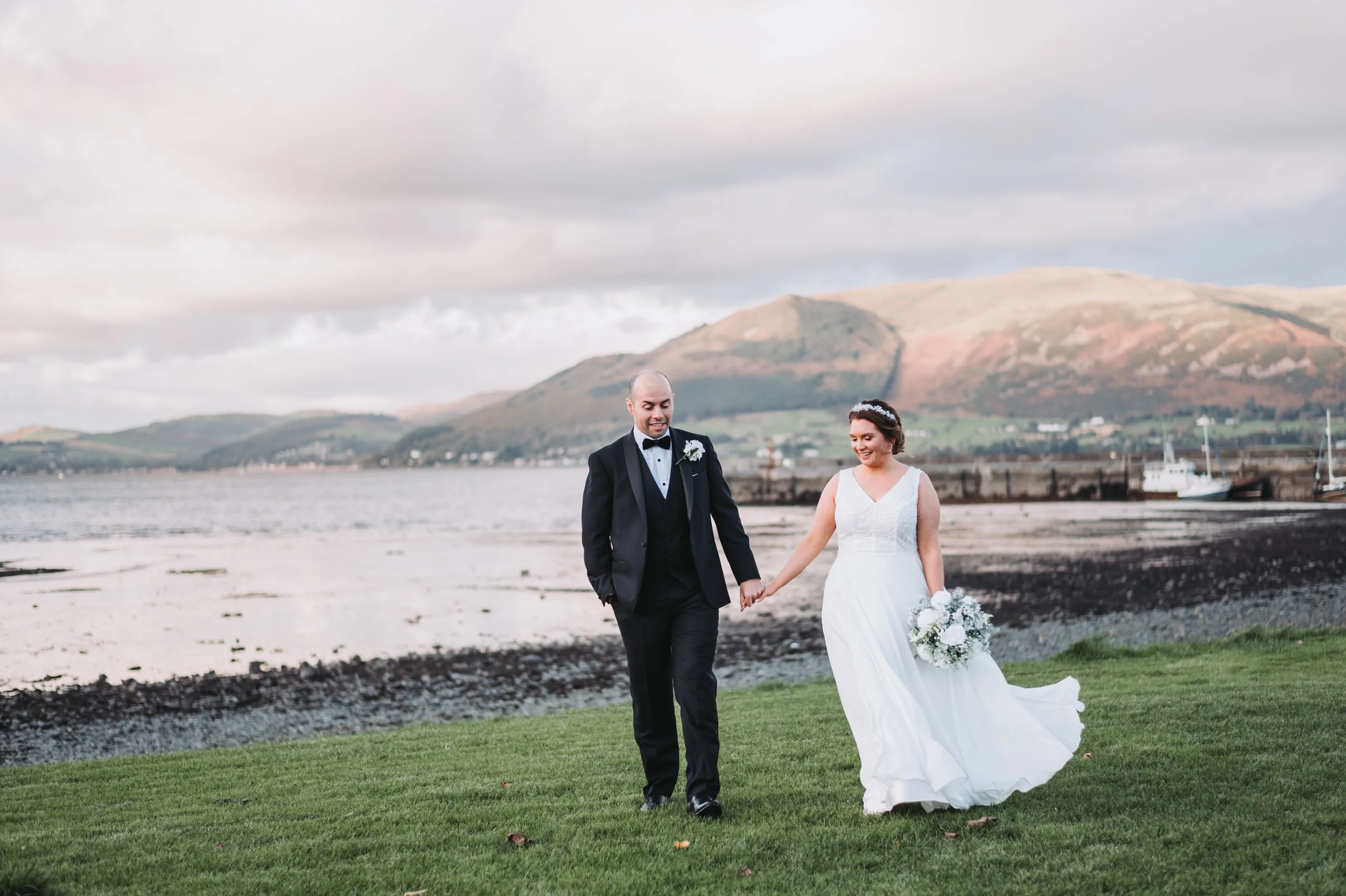 A bride and groom walking hand in hand by a lakeside with mountains in the background, dressed in wedding attire. The bride is holding a bouquet.