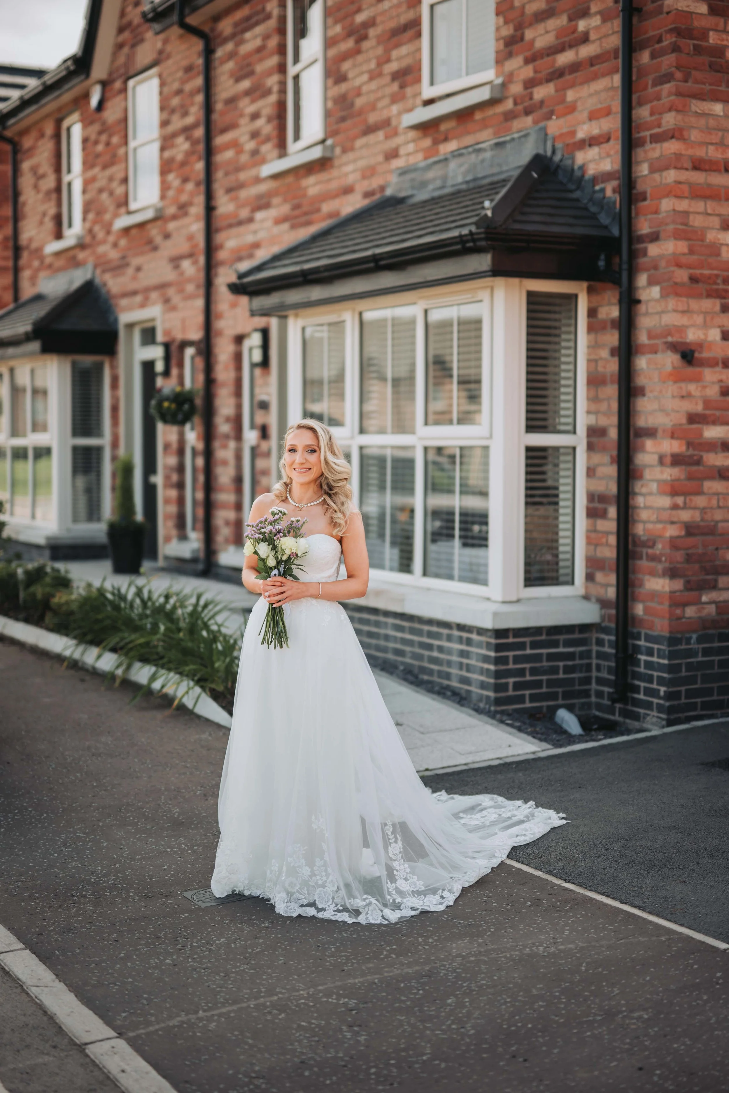 A woman in a white wedding dress holding a bouquet of flowers standing on a sidewalk outside a brick house.