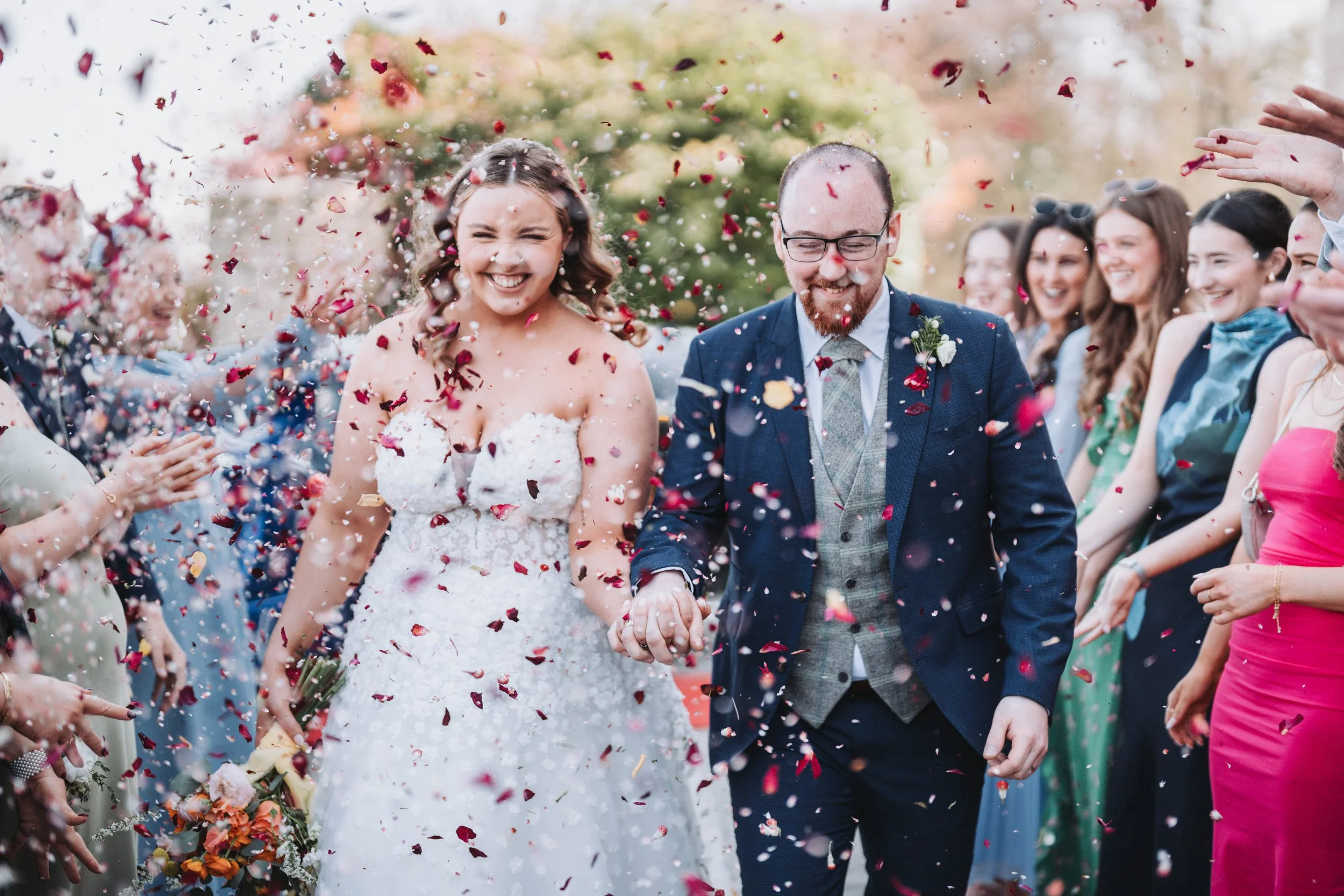 A bride and groom smiling and holding hands as they walk through a shower of confetti, surrounded by happy wedding guests.