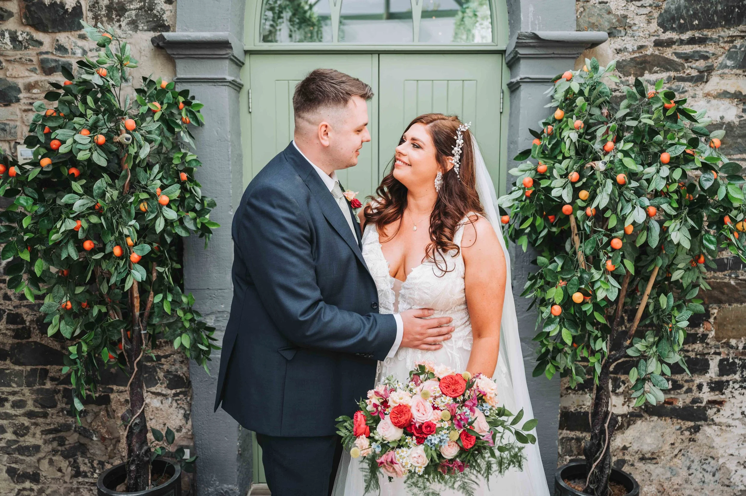 A bride and groom gazing at each other, standing between two small trees with orange fruits, against a stone wall and green door.