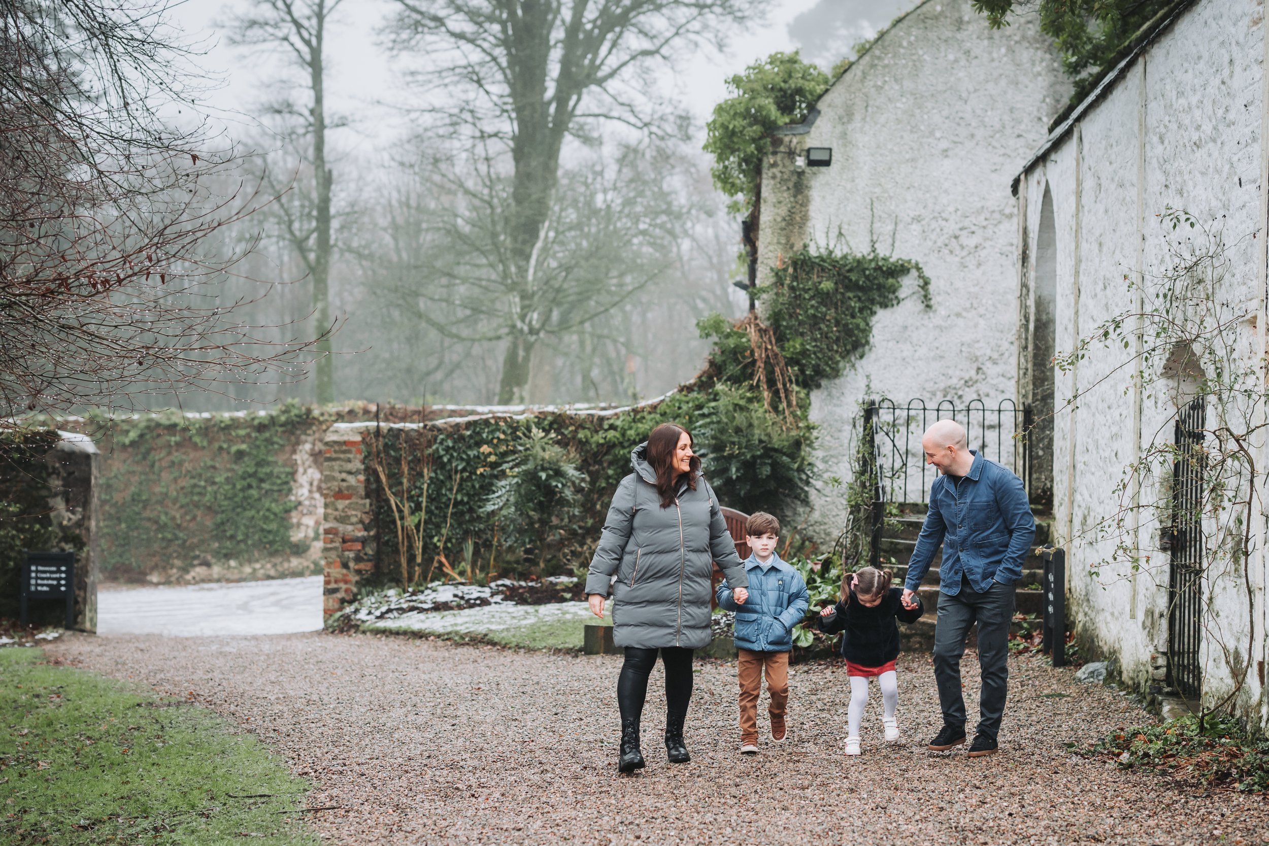 Family walking outside on a gravel path in a courtyard with a white stone building and leafless trees in the background on a foggy day.