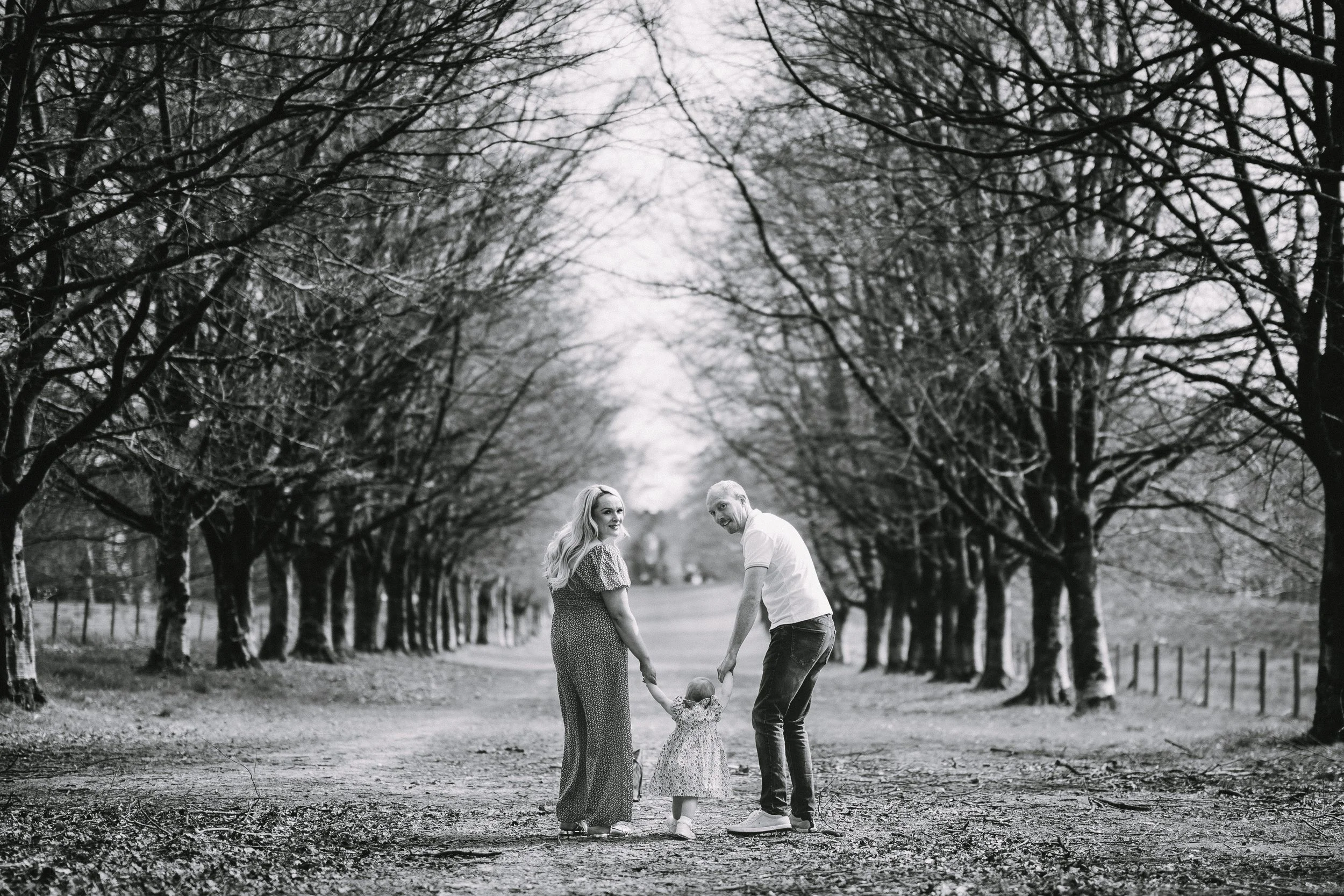 A black and white photo of a family of three walking hand in hand on a tree-lined dirt path in a park or forest, with the parents on either side of their young daughter, who is holding both their hands.