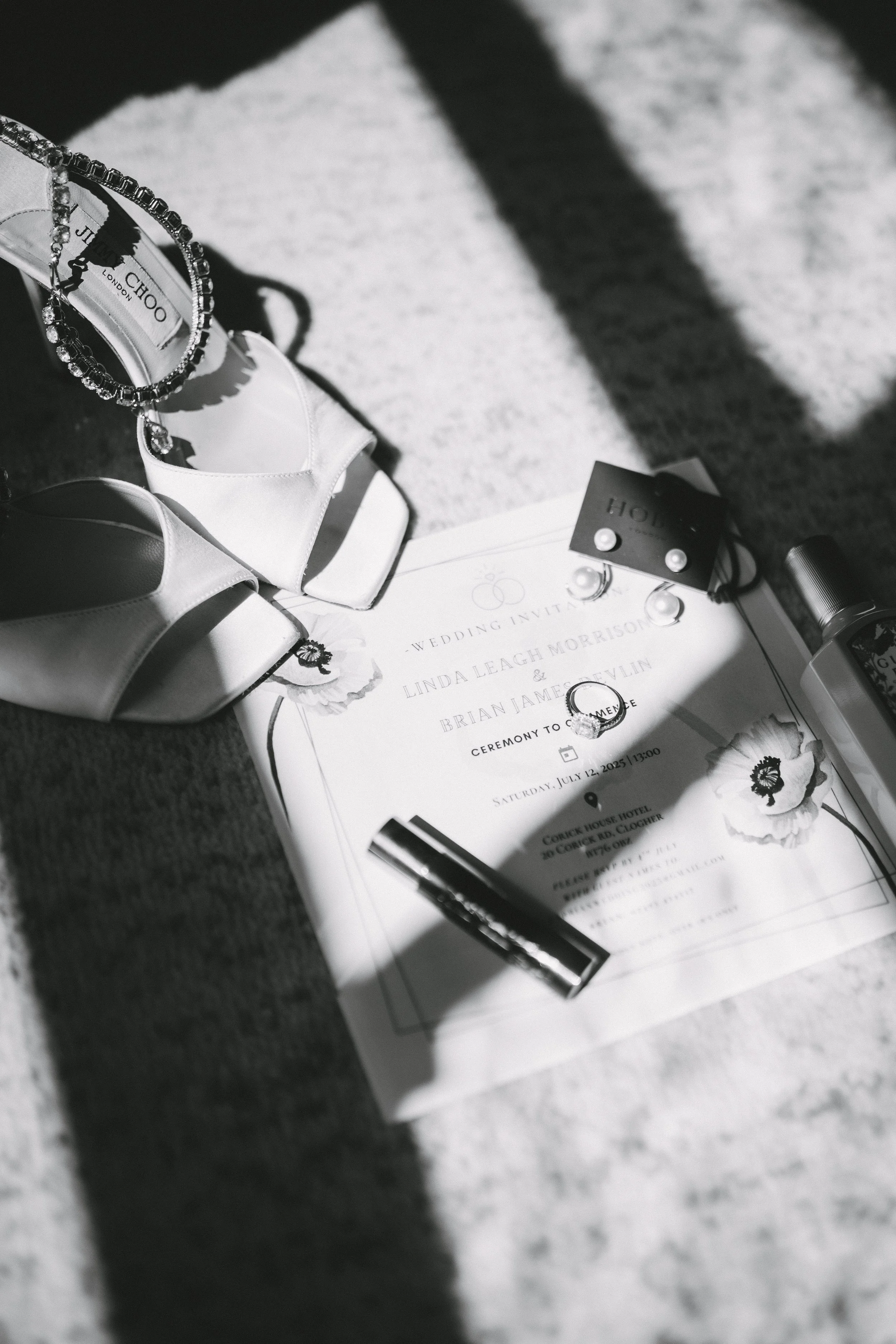 Black and white photo of a wedding invitation surrounded by jewelry, shoes, lipstick, perfume, and a small flower, with shadows cast across the items.