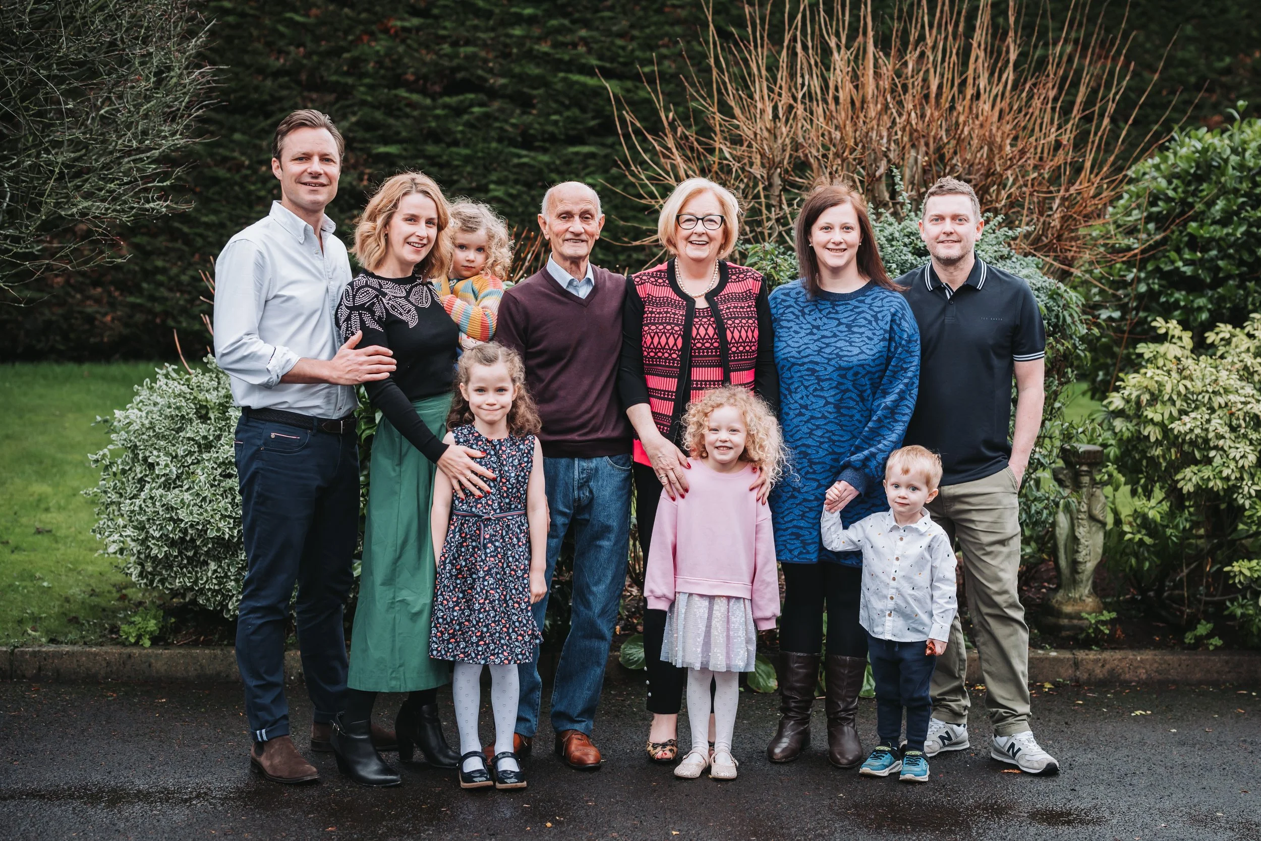 A multigenerational family of ten, including children, adults, and elderly, standing outdoors in a garden with green shrubs and trees in the background, smiling at the camera.