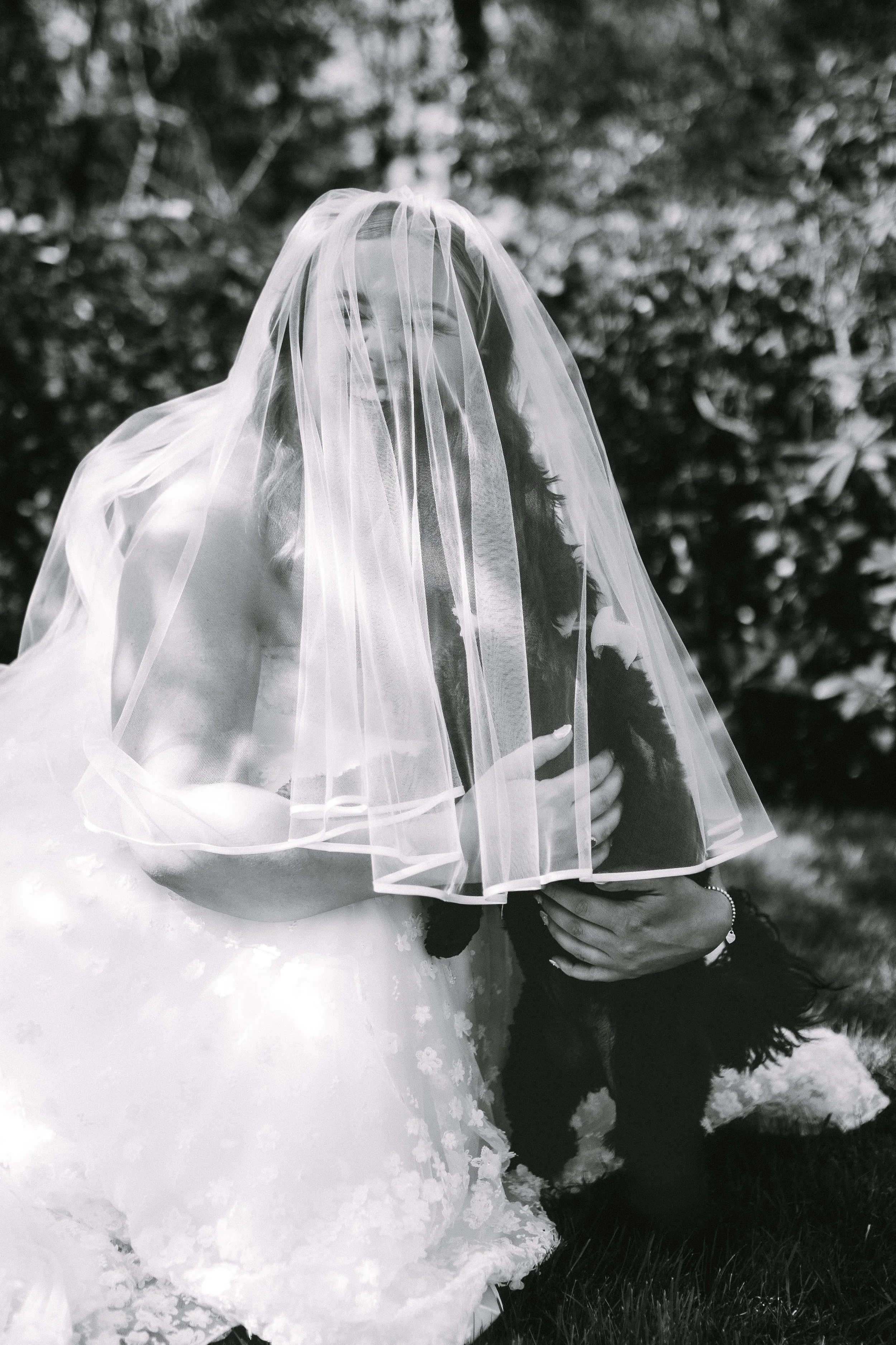 Black and white photo of a woman in a wedding dress, with a veil on her face, hugging a dog outdoors.