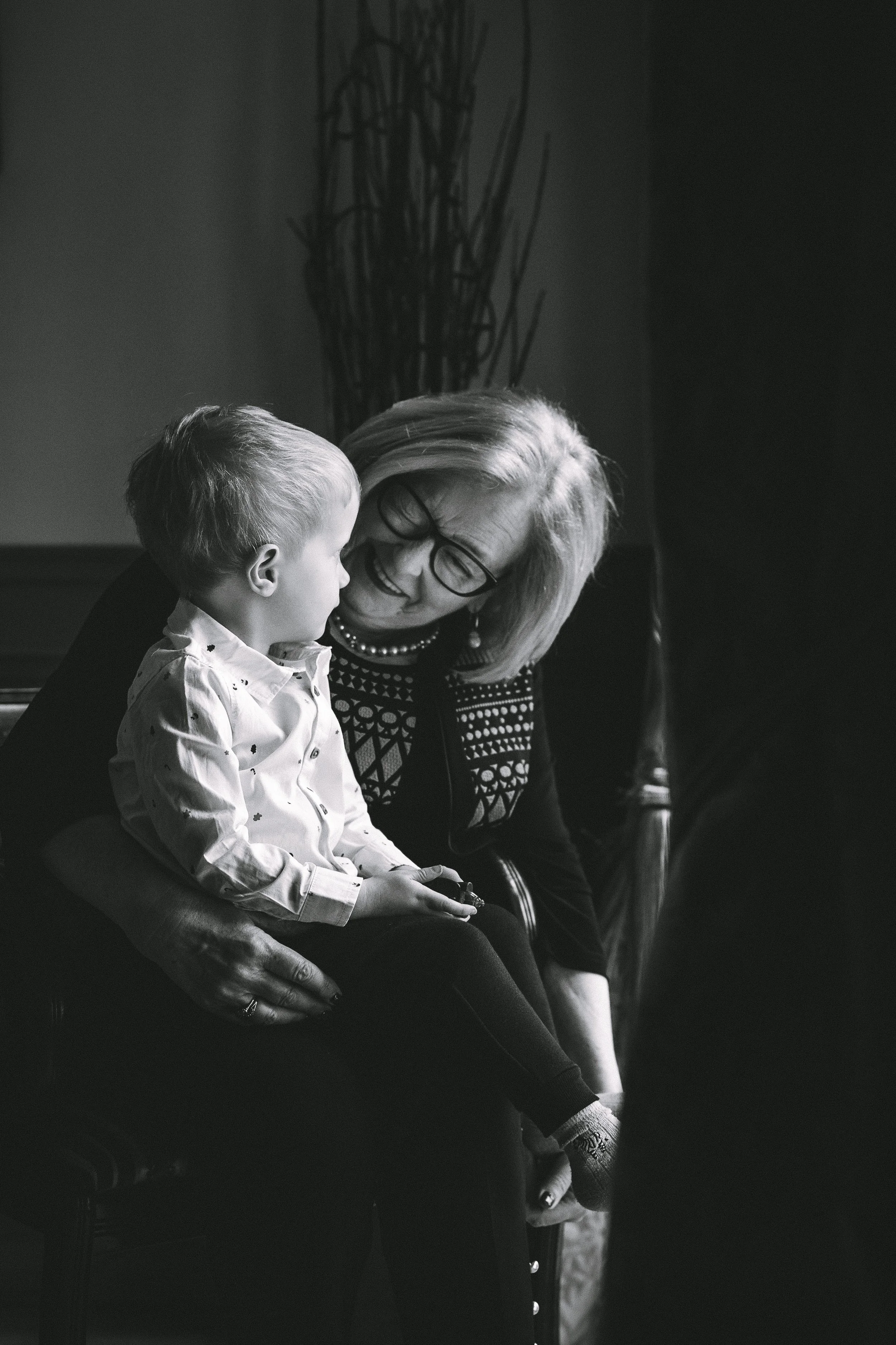 A black-and-white photo of a woman with glasses and a young boy sitting on her lap, sharing a happy moment inside a cozy room.