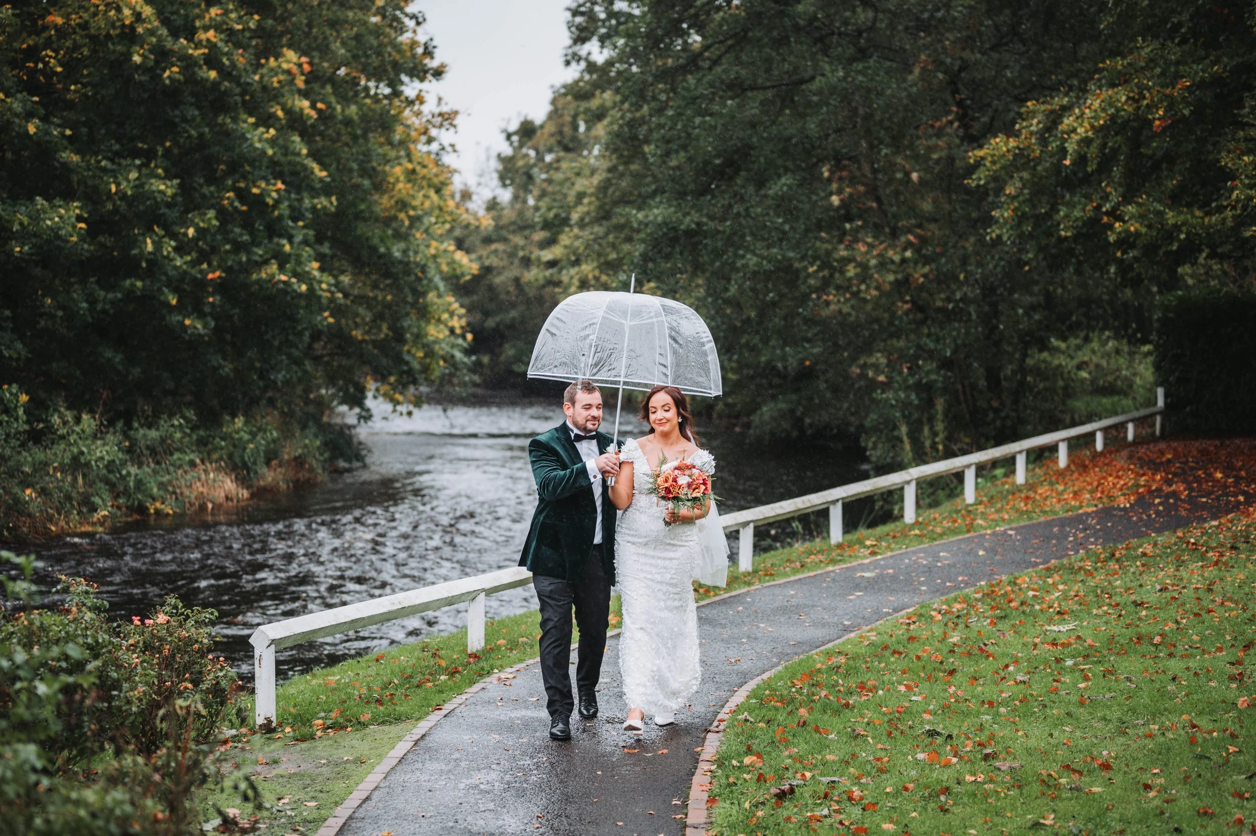A bride and groom walking under a clear umbrella along a park path beside a river on a rainy day, with autumn leaves scattered on the ground.