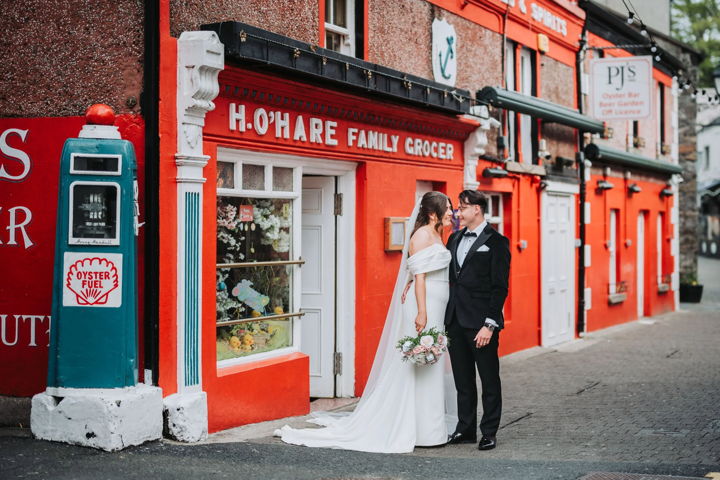 Bride and groom in wedding attire standing close together outside a red building with white trim, holding a bouquet of flowers, smiling at each other.