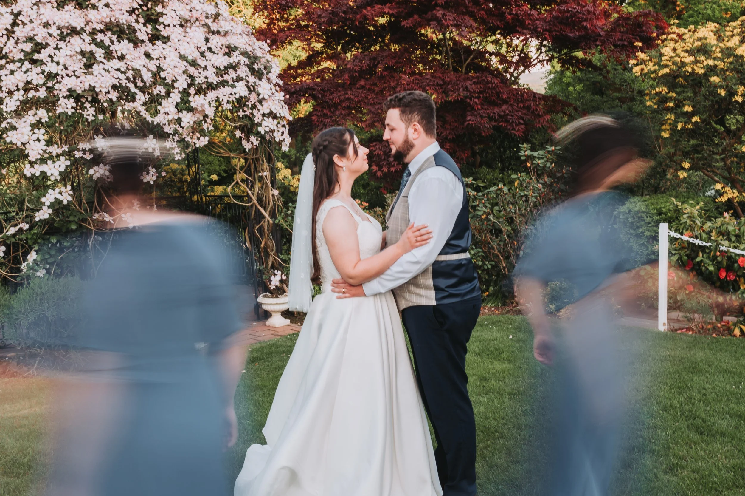 Bride and groom holding each other, facing each other, in a garden with trees and flowers in the background, during a wedding ceremony.