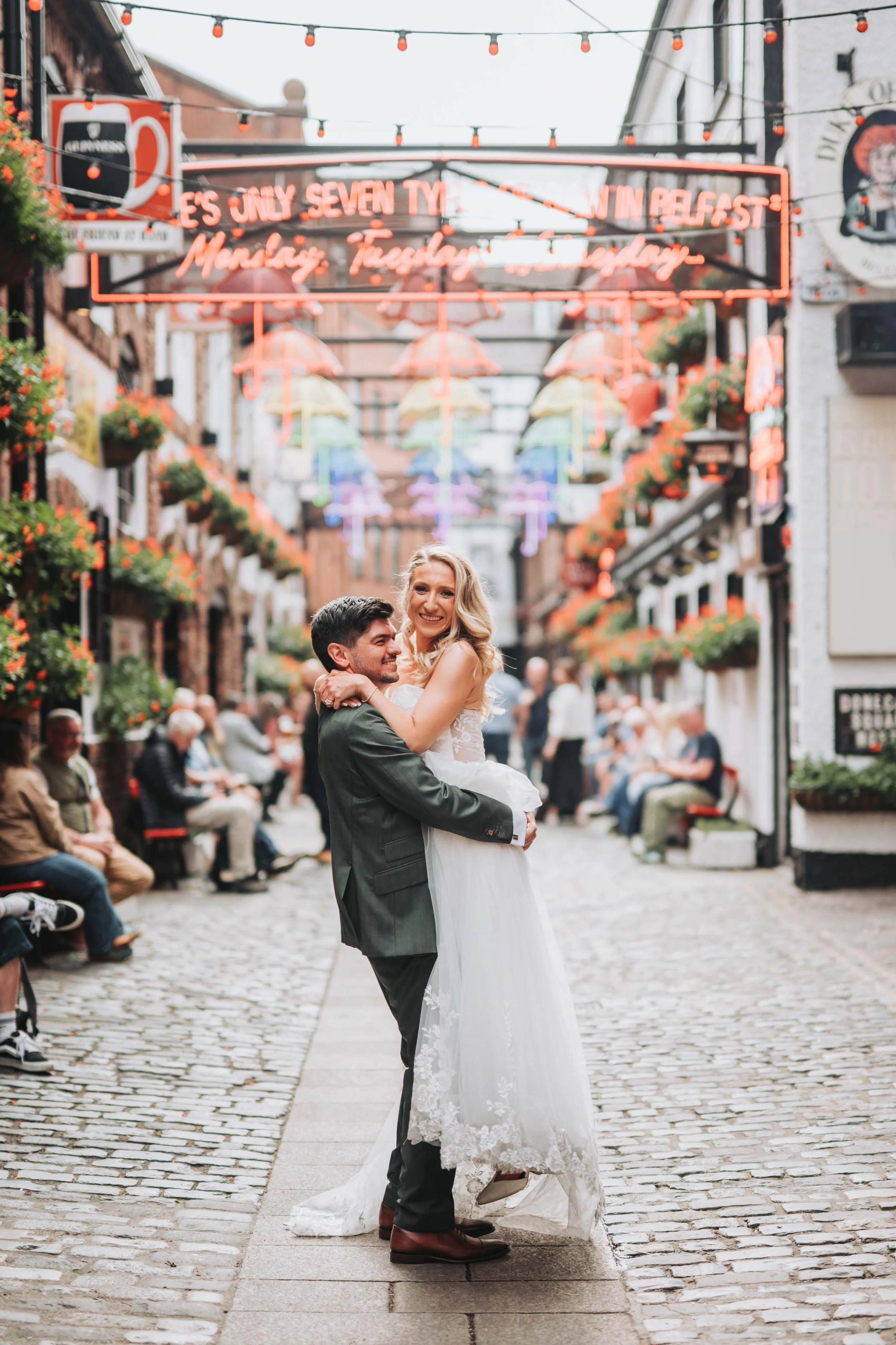 A newlywed couple, the groom in a dark suit and the bride in a white wedding dress, embracing and smiling on a cobblestone street decorated with colorful umbrellas hanging overhead, with people seated on benches and dining establishments in the backg
