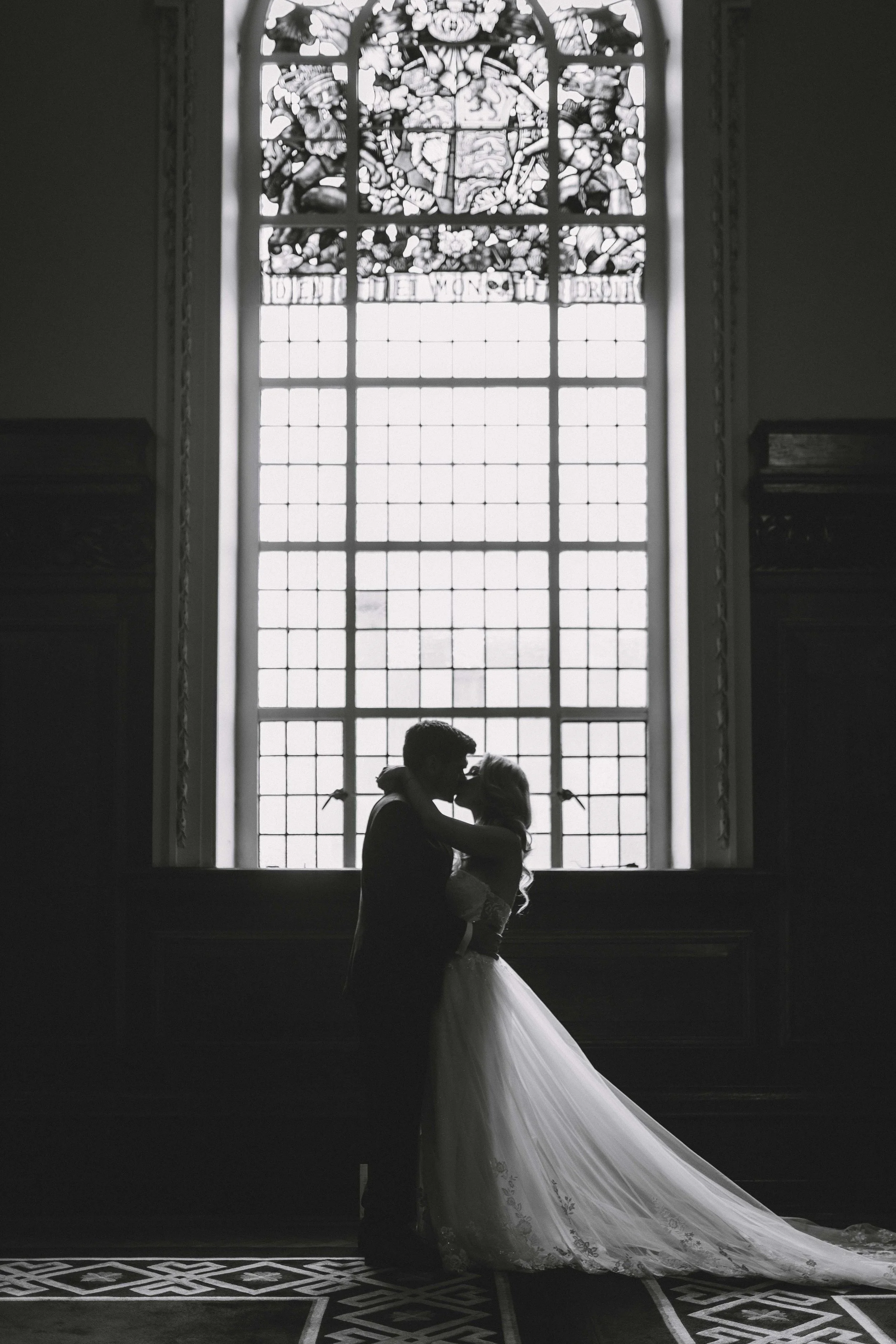 Silhouette of a bride and groom sharing a kiss in front of a large stained glass window in a church.