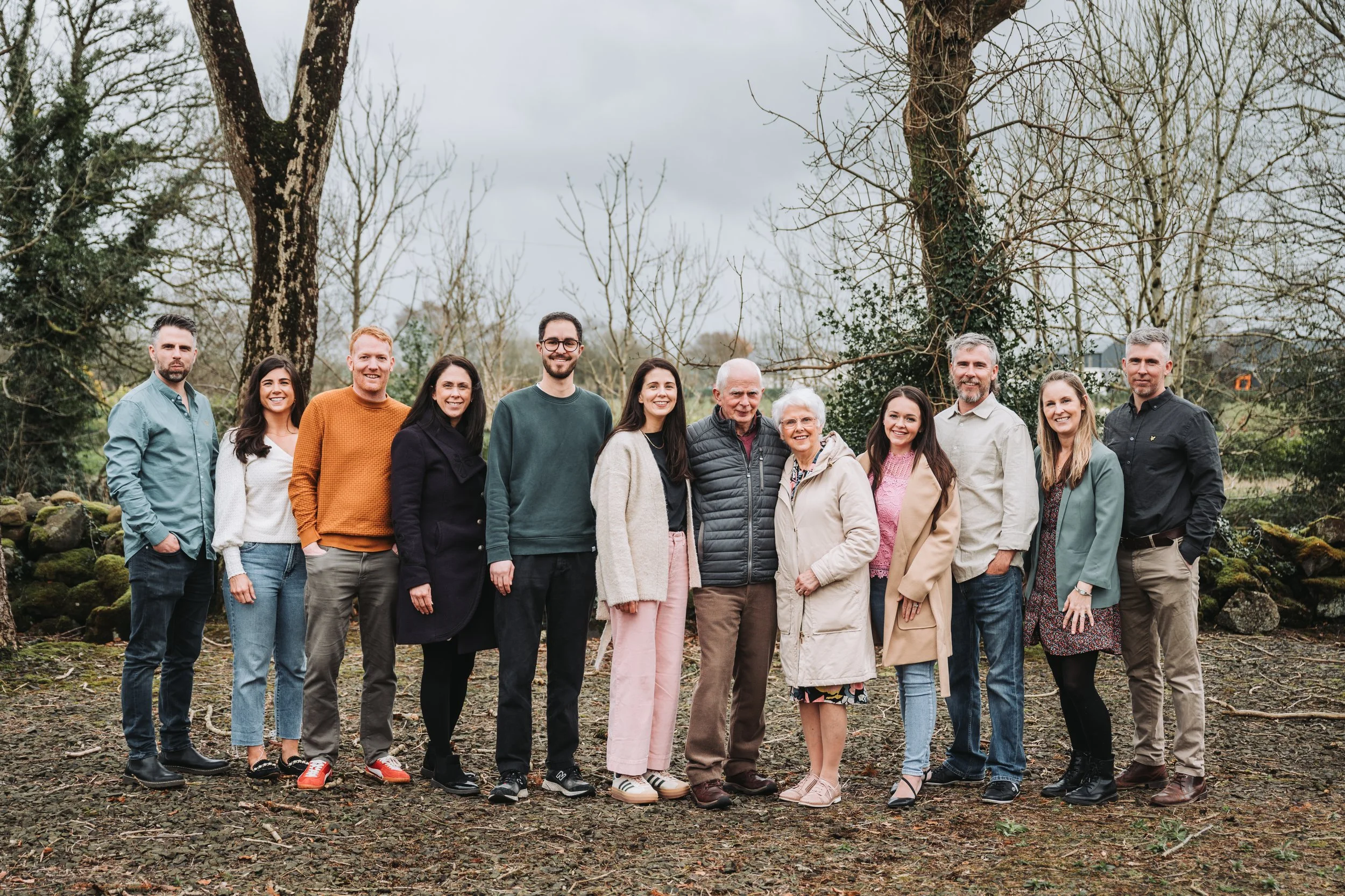 Group of fifteen people standing outdoors on a cloudy day, smiling, with trees and rocks in the background.