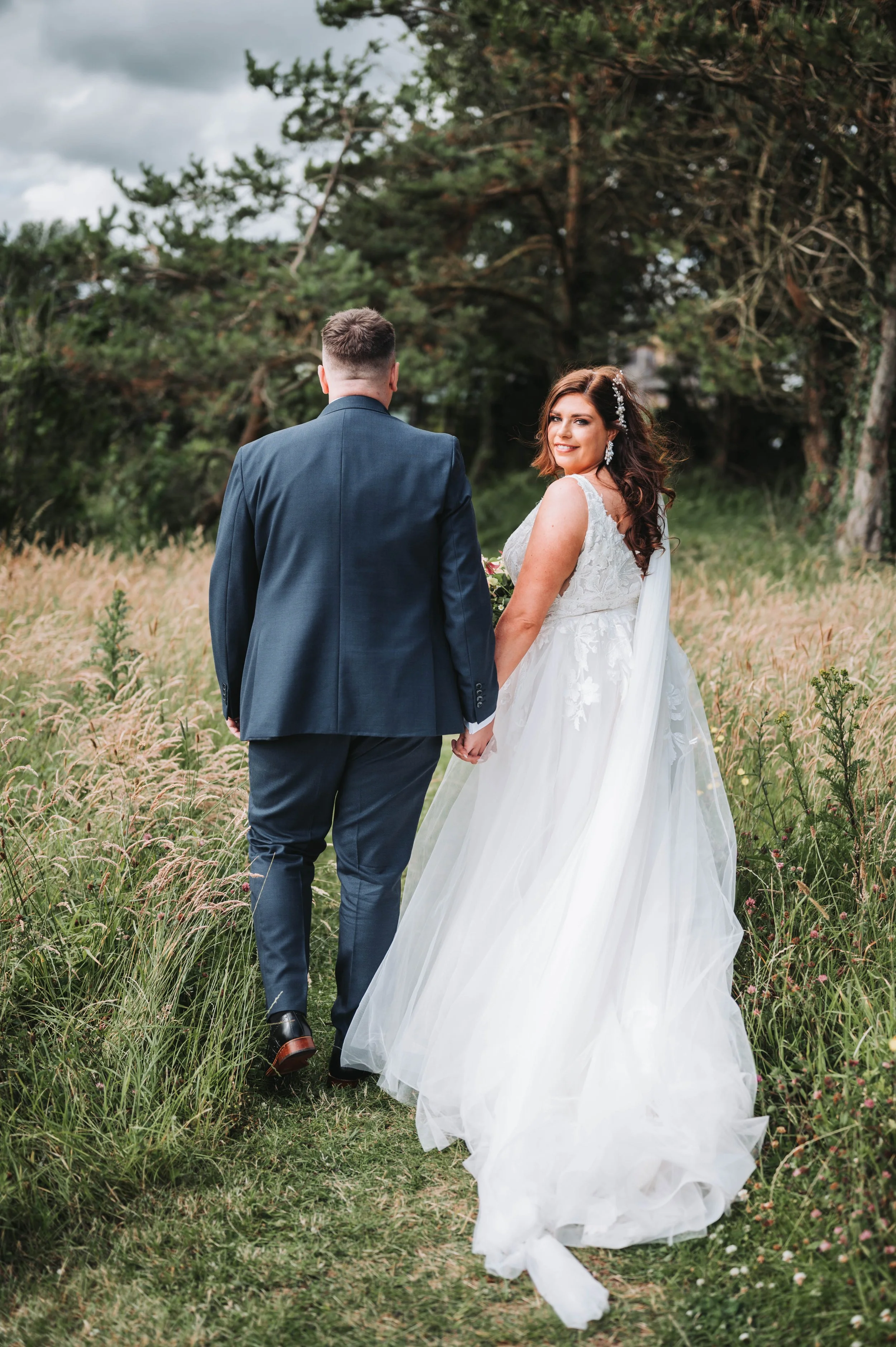 A newlywed couple walking hand in hand on a grassy trail surrounded by wildflowers, with trees and a cloudy sky in the background. The bride is wearing a white wedding gown and the groom a dark suit.