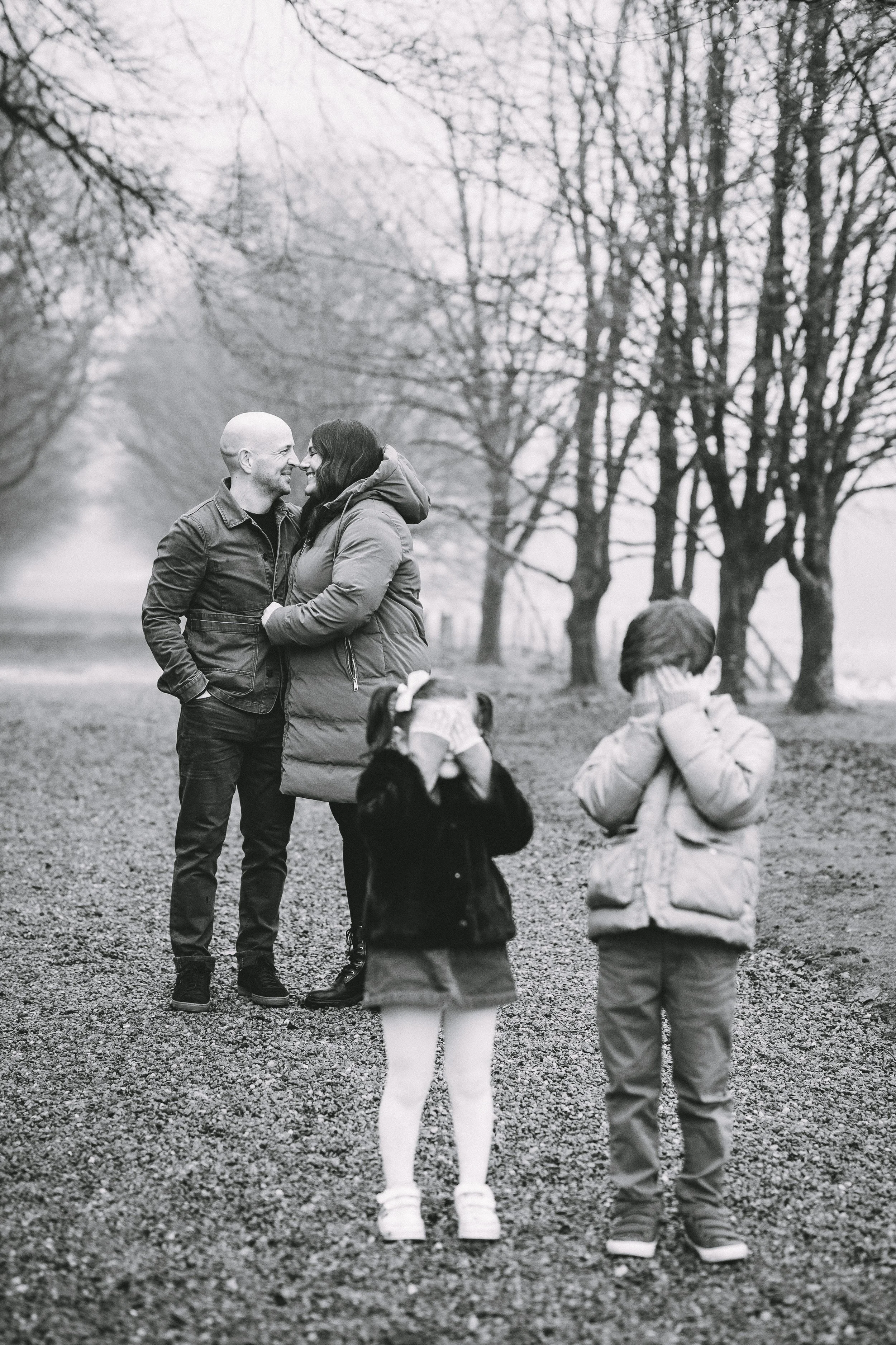A black and white photo of a couple smiling and holding each other in a park, with two children in the foreground covering their eyes.