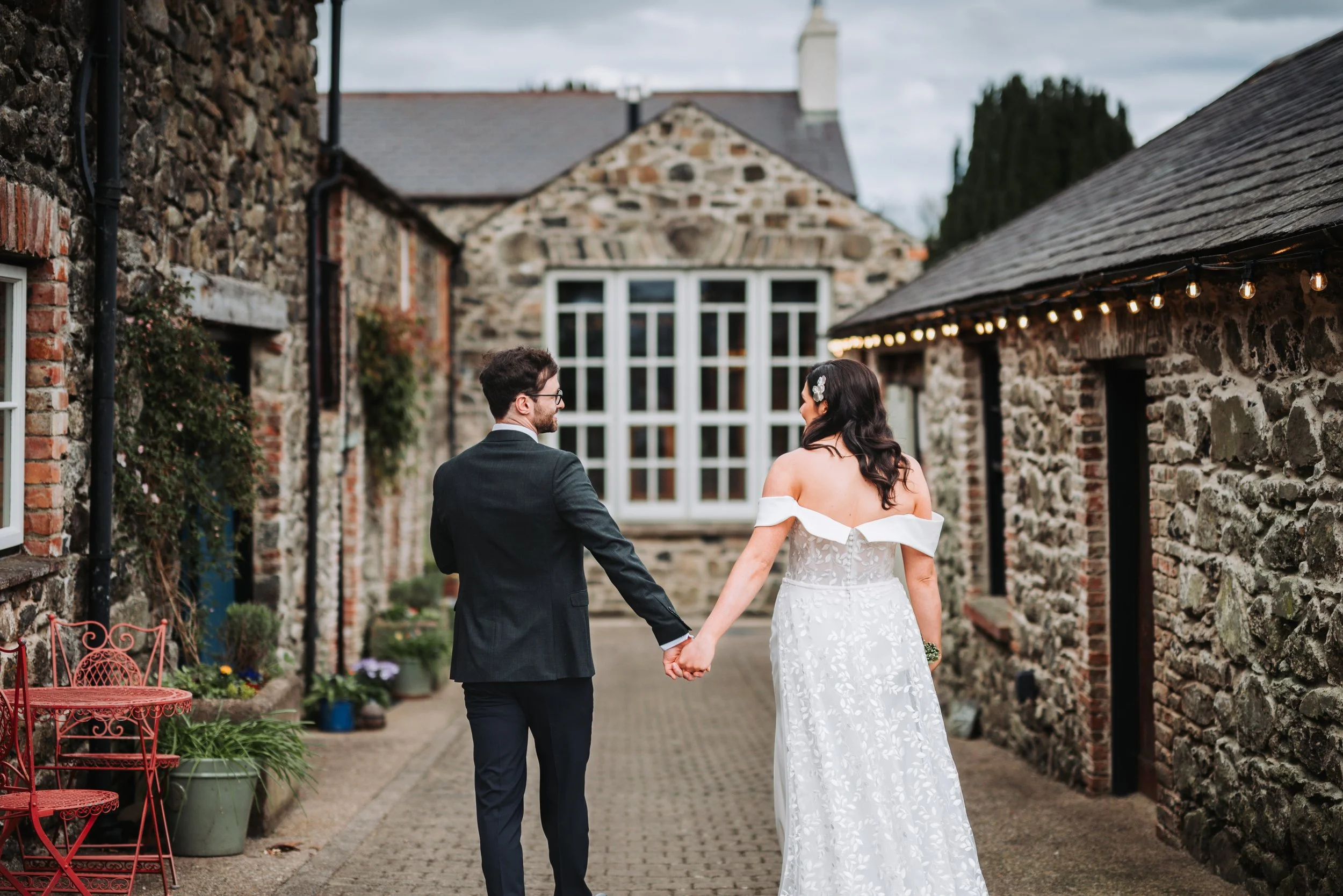 A newlywed couple holding hands walking outdoors between rustic stone buildings with string lights, plants, and outdoor furniture.