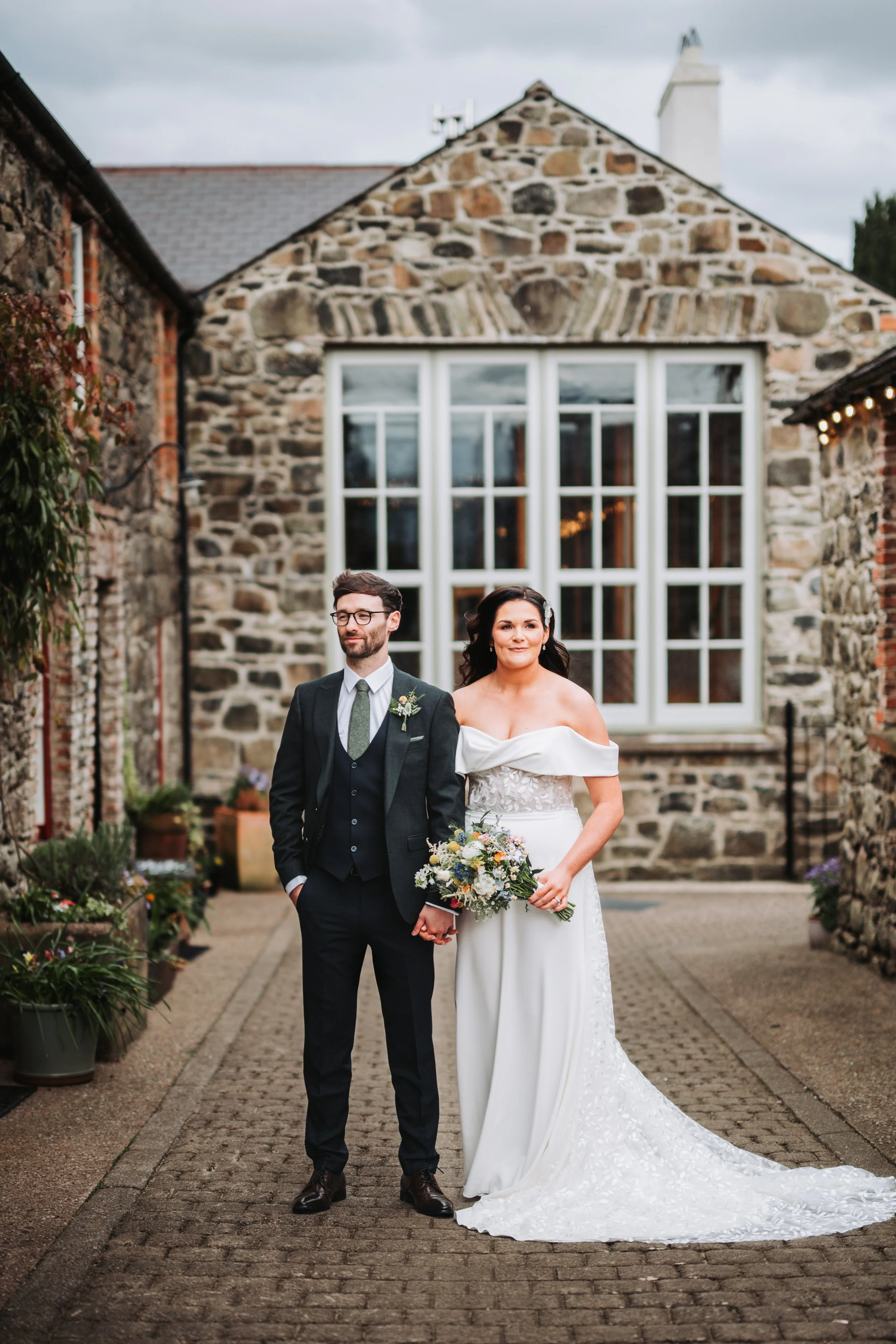 A newlywed couple standing outside in a narrow brick-paved alley, holding hands, with a stone building and large window behind them. The bride is in a white off-the-shoulder wedding dress holding a bouquet, and the groom is in a dark suit with a bout