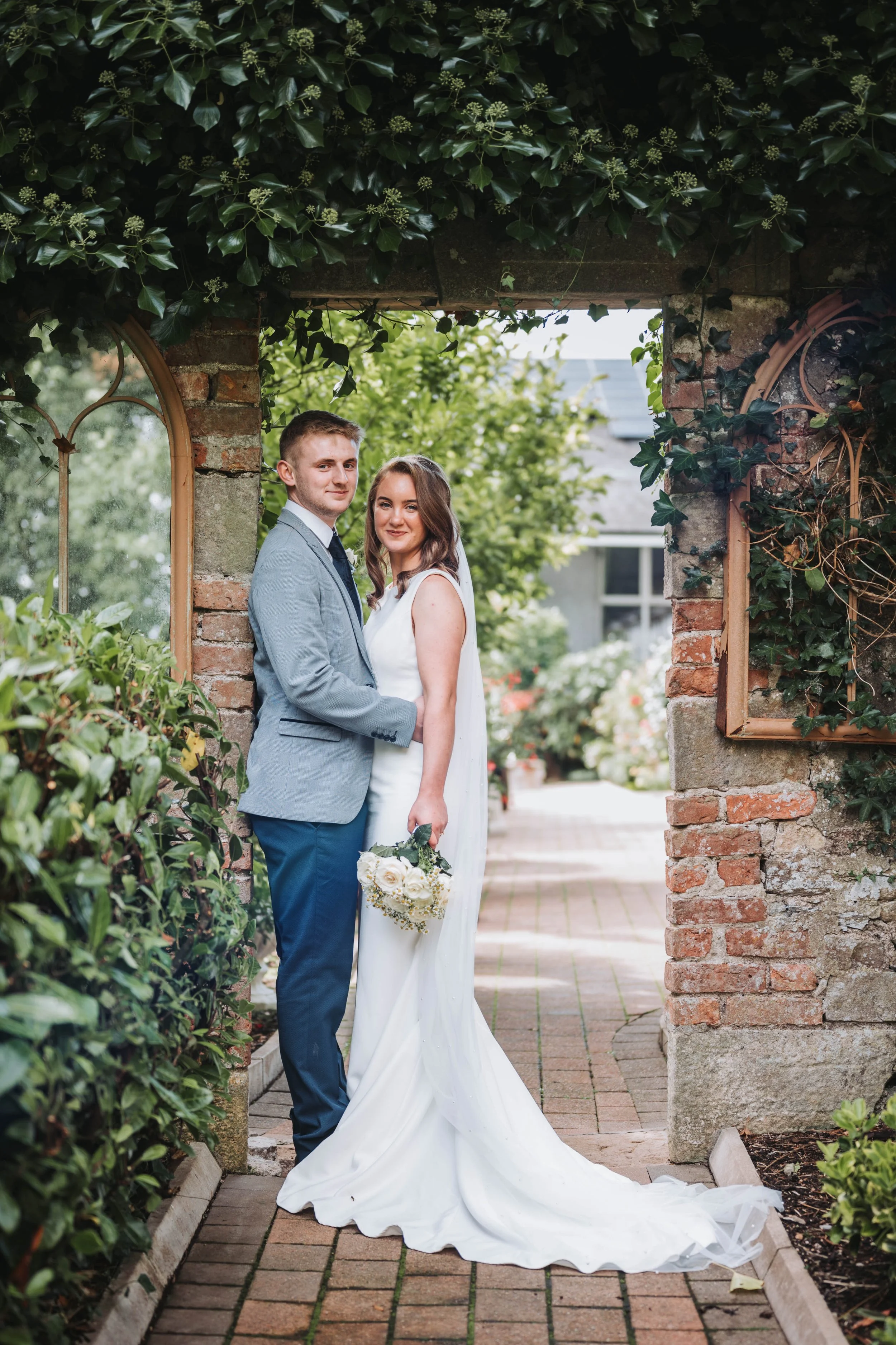 A bride and groom standing together in a garden archway, surrounded by greenery and brick walls, on their wedding day.