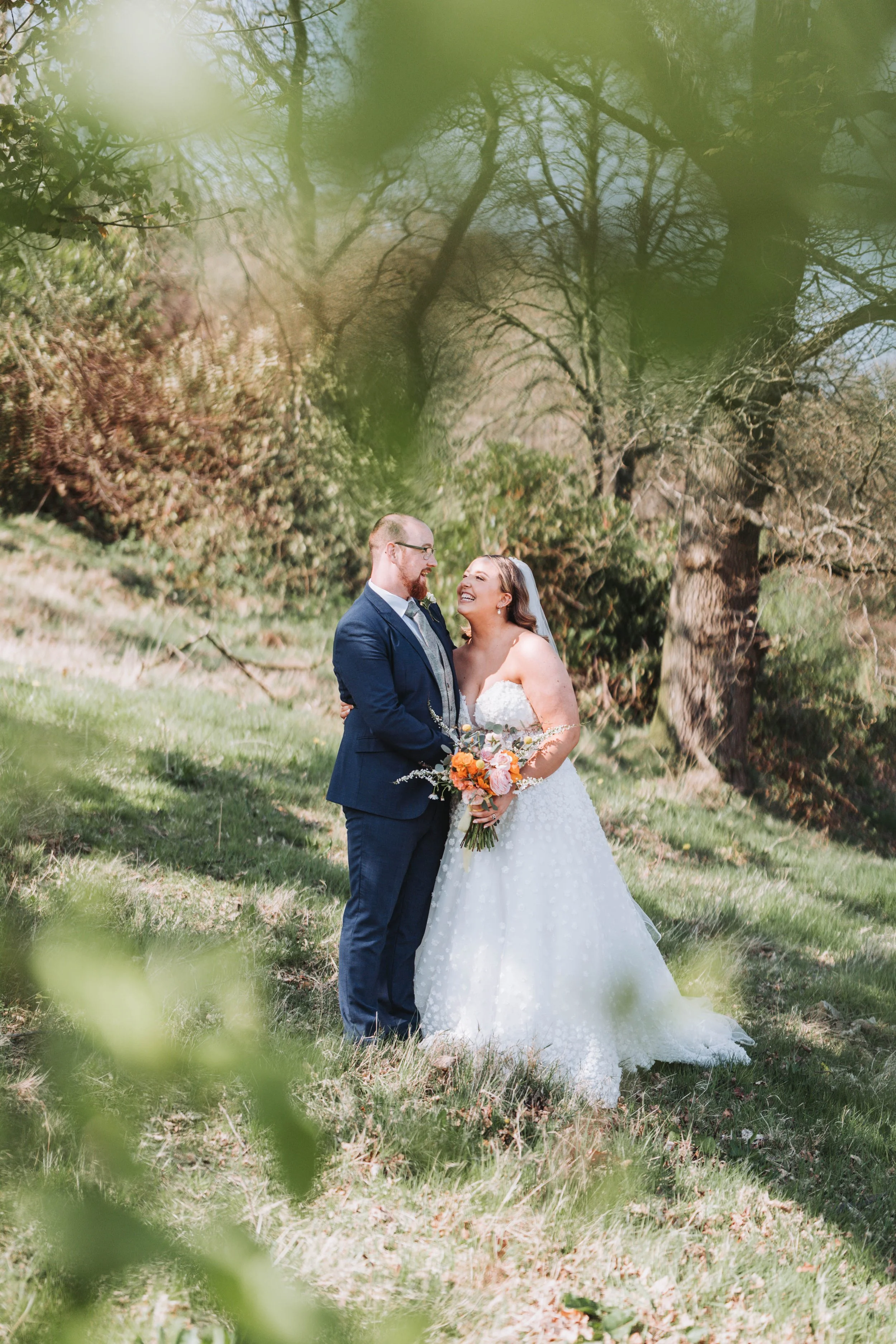 A bride and groom standing in a grassy outdoor area, smiling and looking at each other, with trees in the background and green leaves partially obscuring the scene.