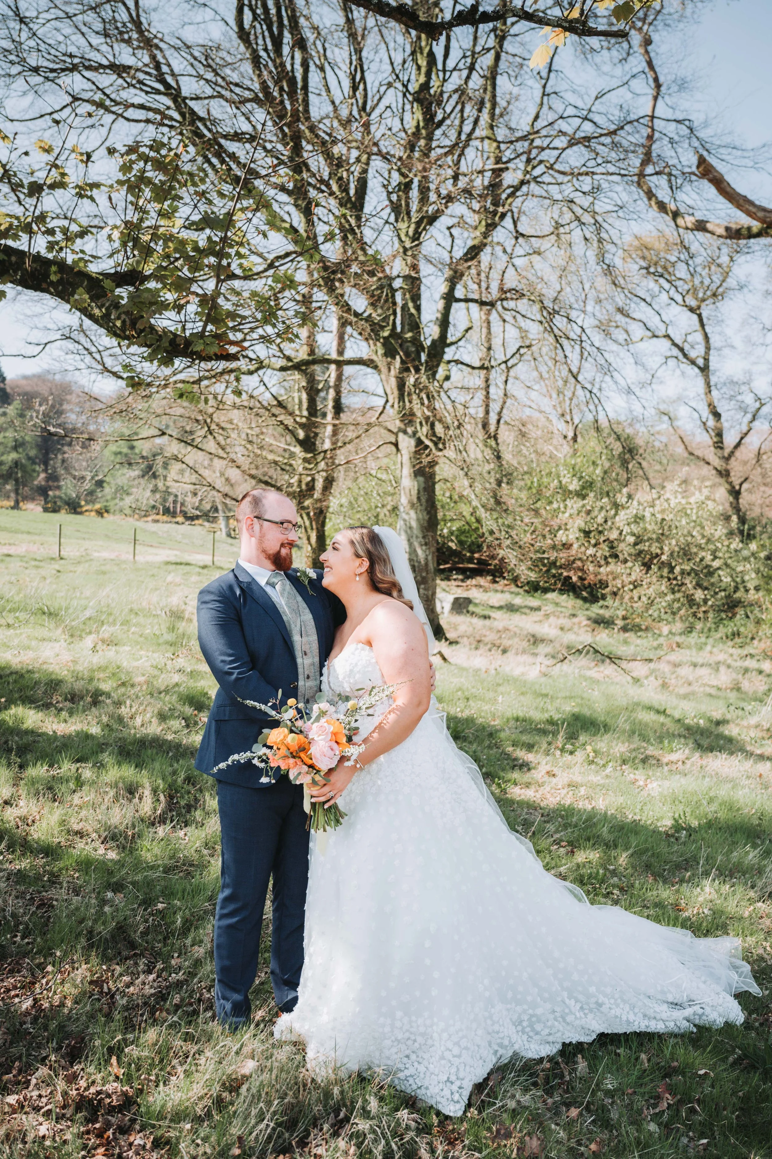 A bride and groom smiling at each other outdoors on a sunny day, standing on grass with a large tree and a partly cloudy sky in the background.