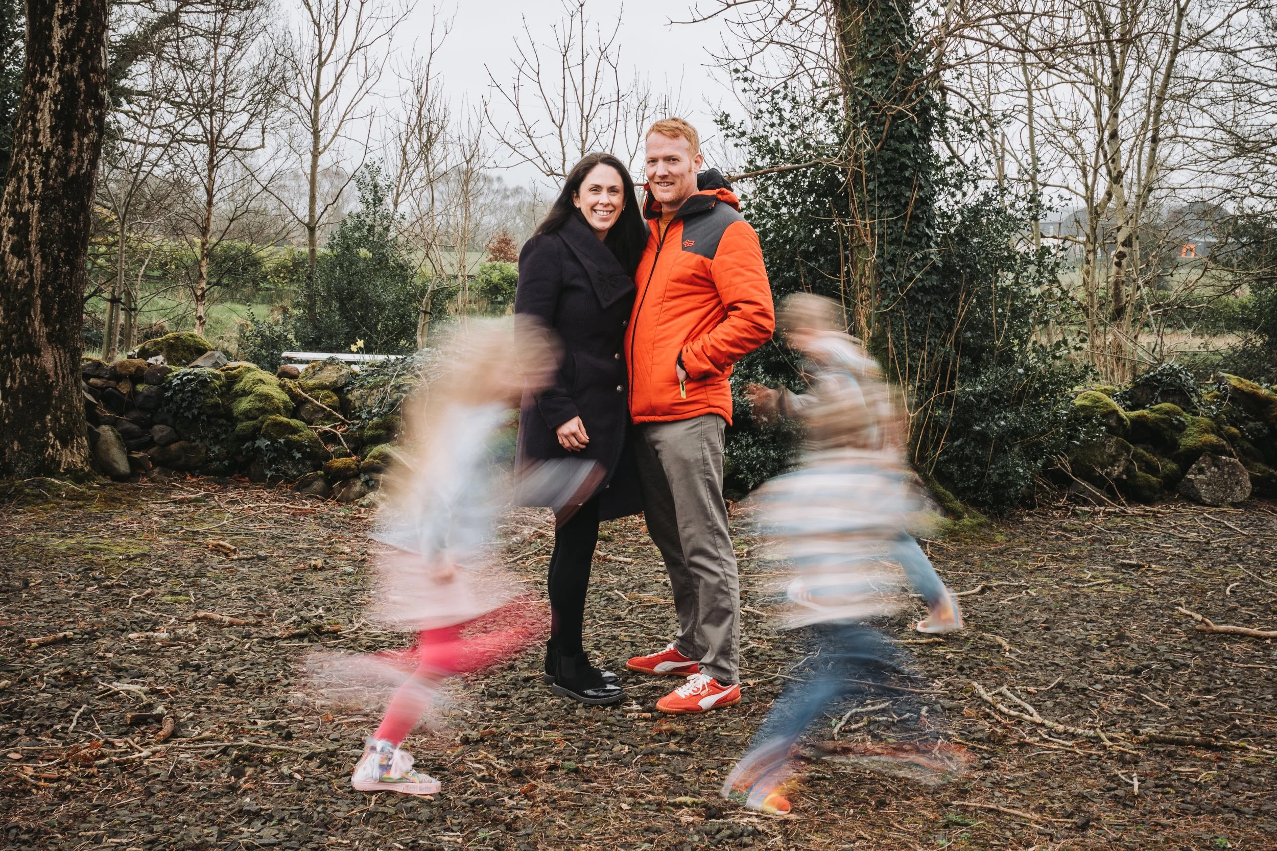 A smiling couple standing outdoors in a park on a cloudy day, with two blurred children running around them.