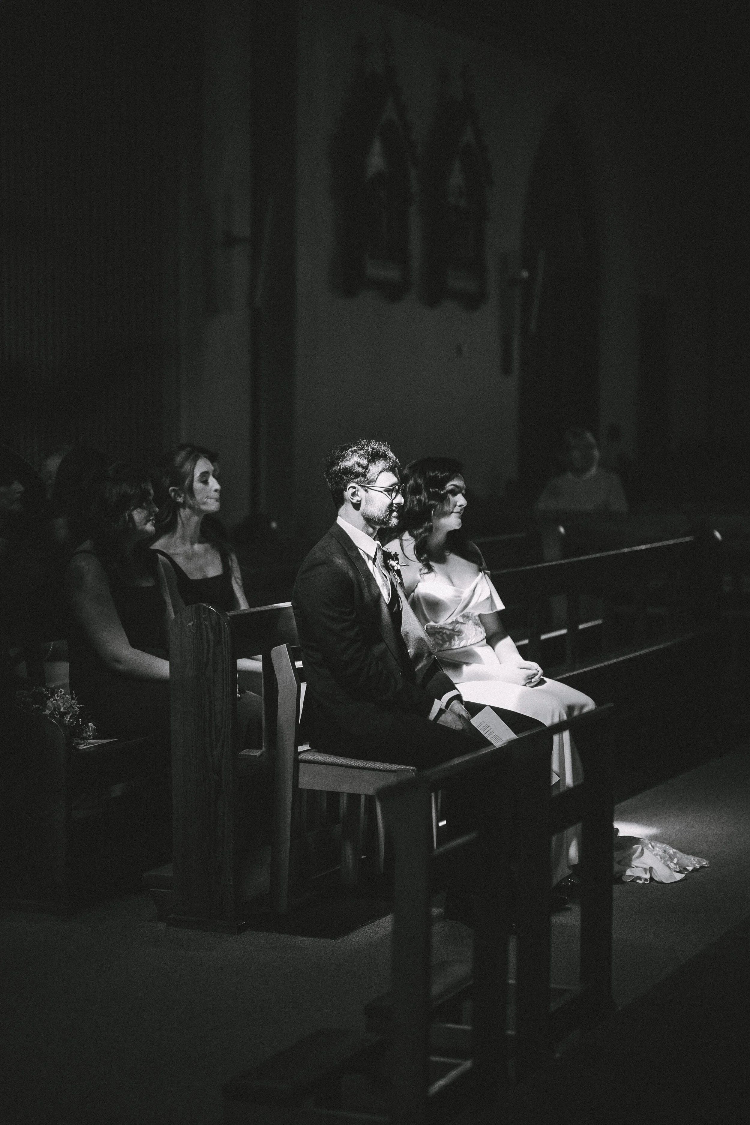 Black and white photograph of a wedding ceremony showing a couple sitting in church pews, with a small group of guests seated behind them, focused on the couple. The couple appears calm and attentive, with the bride in a white gown and the groom in a