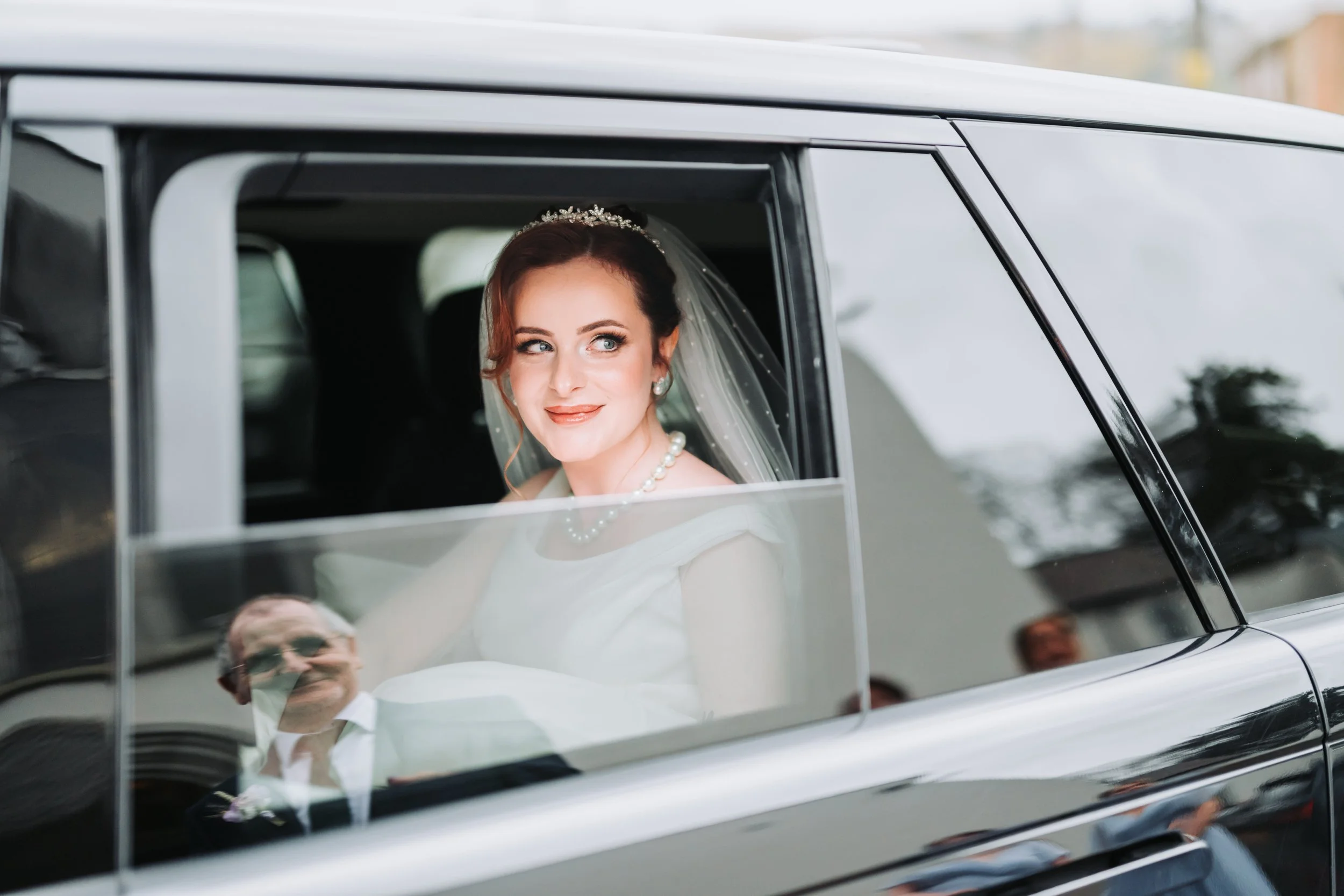 Bride with makeup and pearl jewelry sitting in a black car, looking out the window with a slight smile.