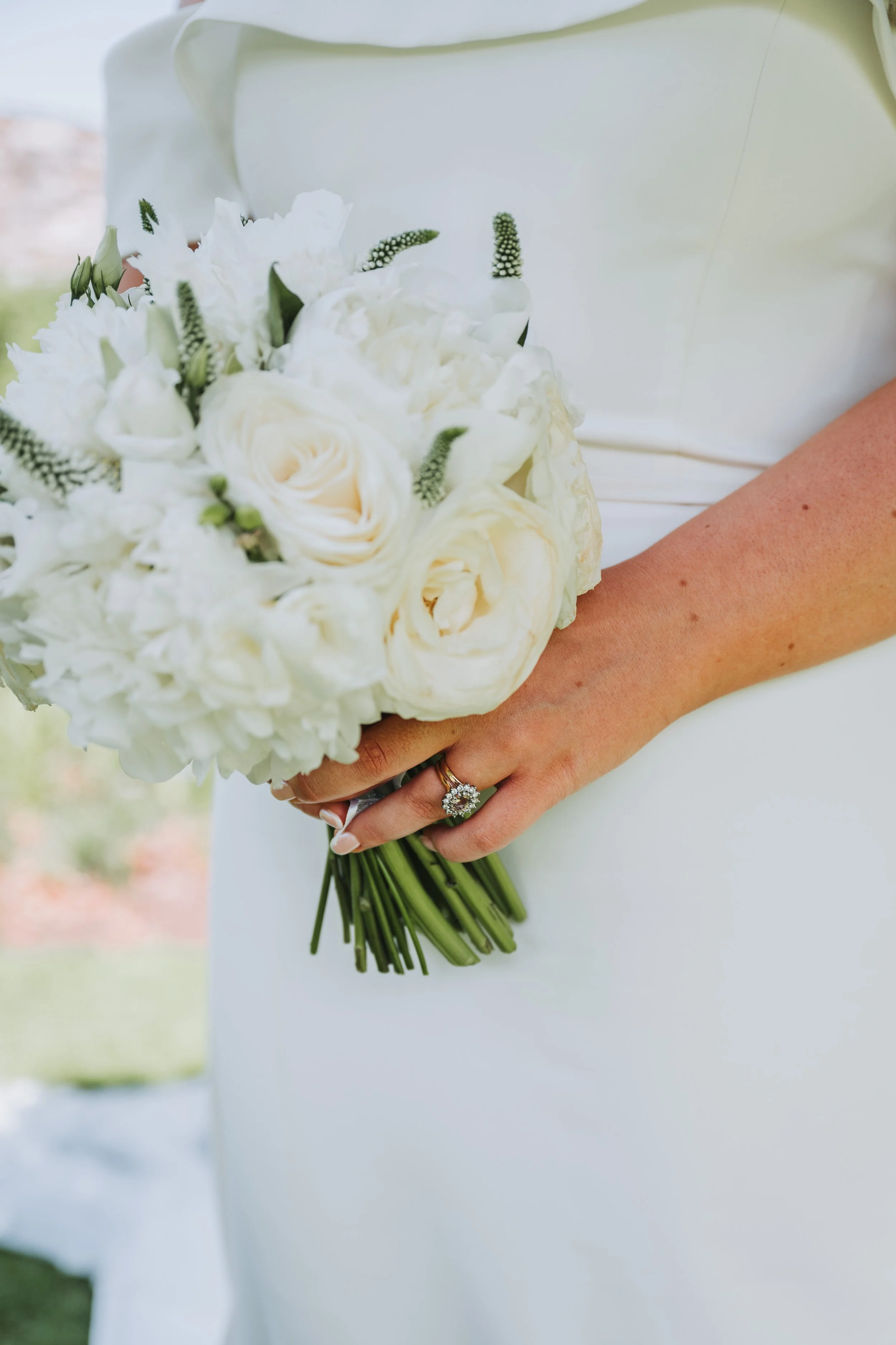 A bride holding a bouquet of white flowers and wearing an engagement ring, dressed in a white wedding gown.