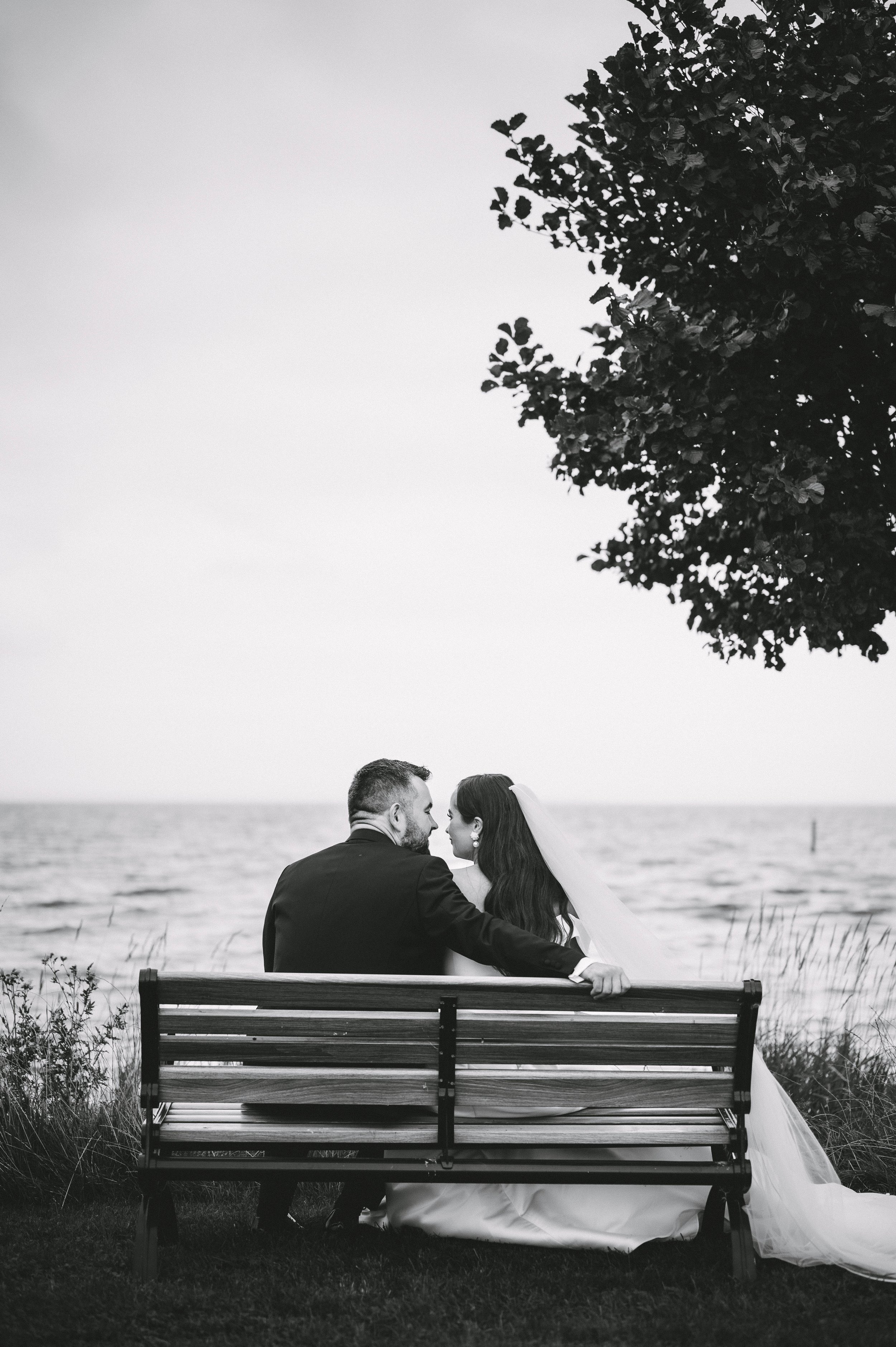 Black and white photograph of a newlywed couple sitting on a park bench by the ocean, sharing an intimate moment with their foreheads touching. The bride is wearing a wedding dress and veil, and the groom is in a suit. A tree hangs over them on the r