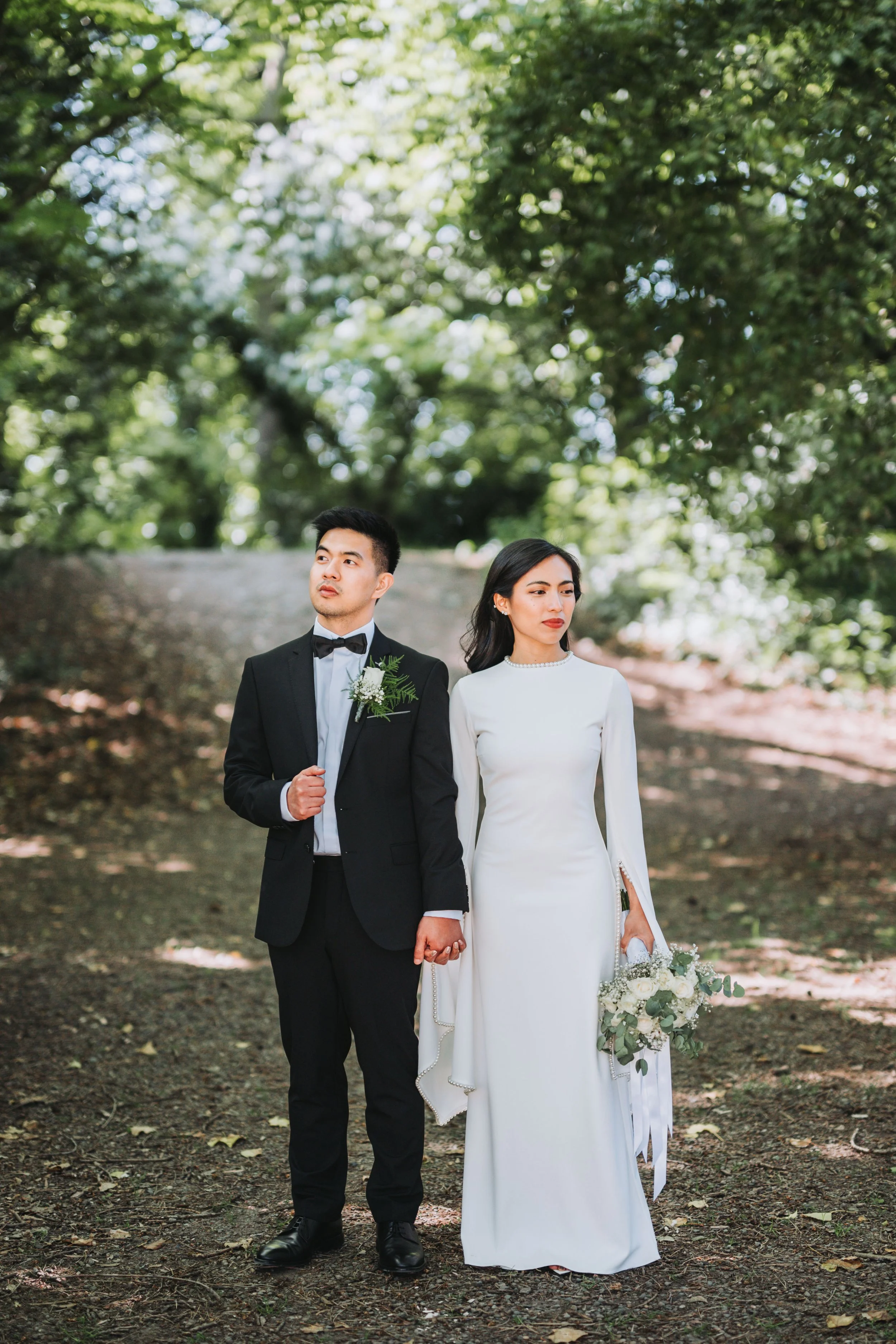 A couple dressed in wedding attire standing outdoors on a dirt path surrounded by trees. The man is in a black tuxedo with a bow tie, holding his jacket lapel, while the woman is in a white wedding gown holding a bouquet of white flowers and greenery