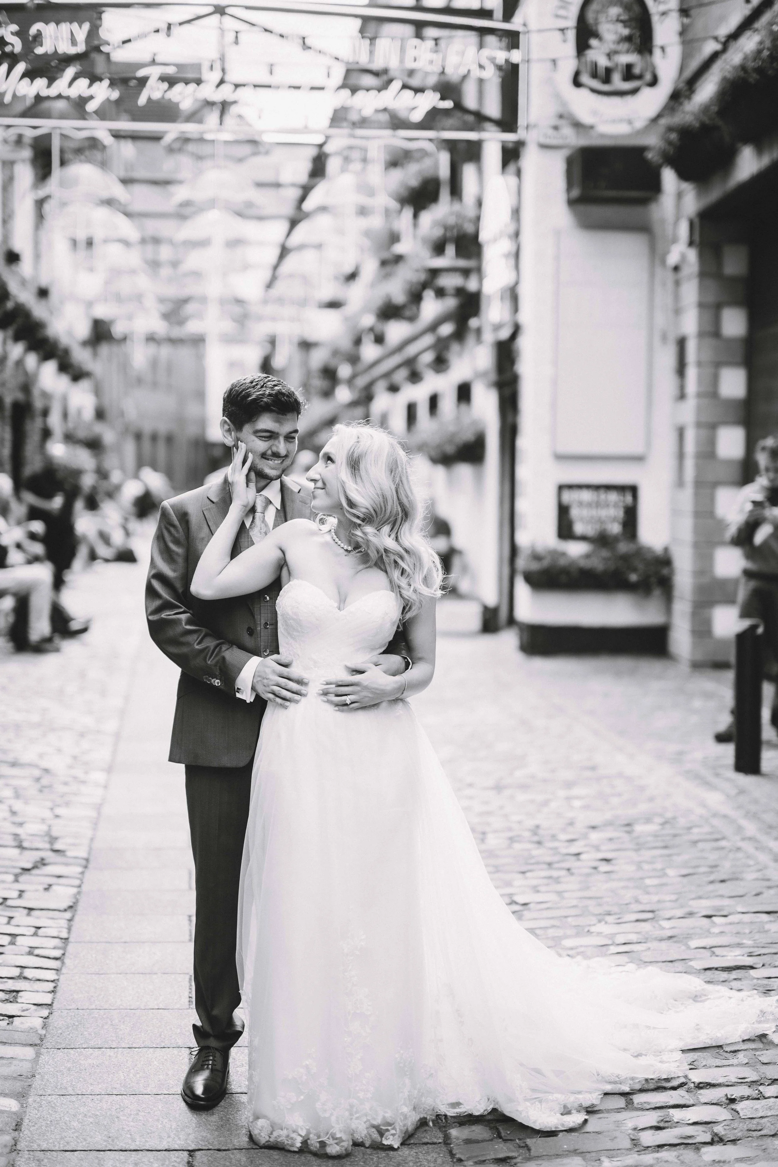 A black-and-white photo of a newlywed couple standing on a cobblestone street, smiling and looking at each other. The bride is wearing a strapless wedding gown, and the groom is in a suit. There are blurred people and storefronts in the background wi