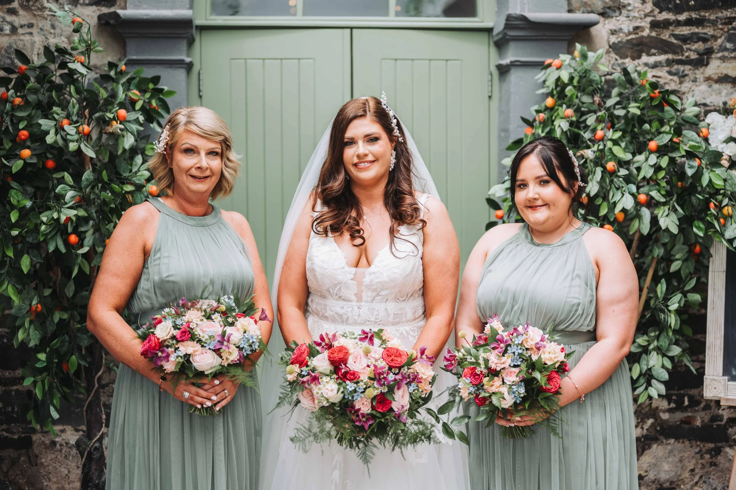A bride in a white wedding dress standing between two bridesmaids in sage green dresses, all holding bouquets of pink, purple, and white flowers, in front of greenery and stone wall.