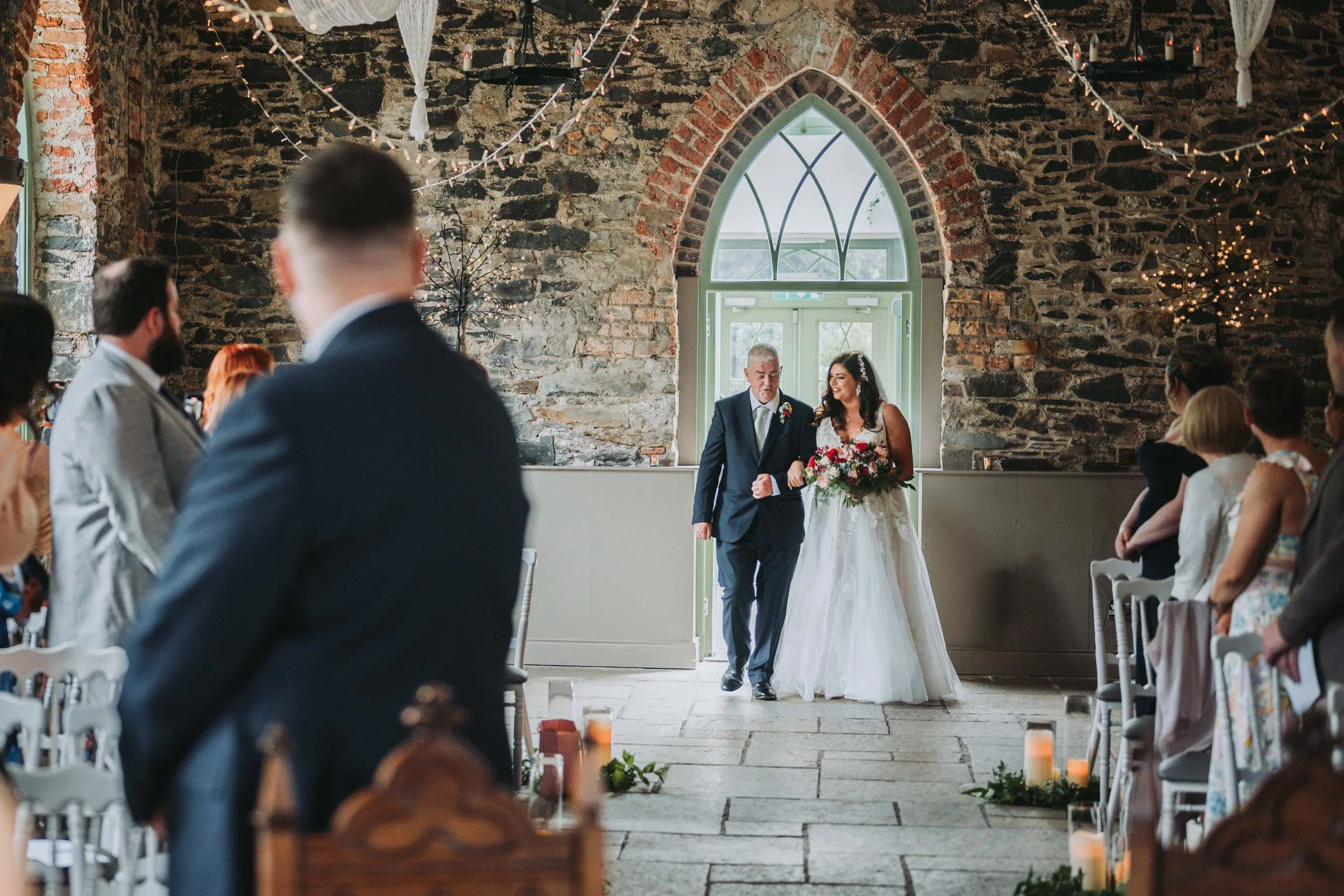 A bride walking down the aisle with an older man in a church with exposed brick walls, while guests watch.